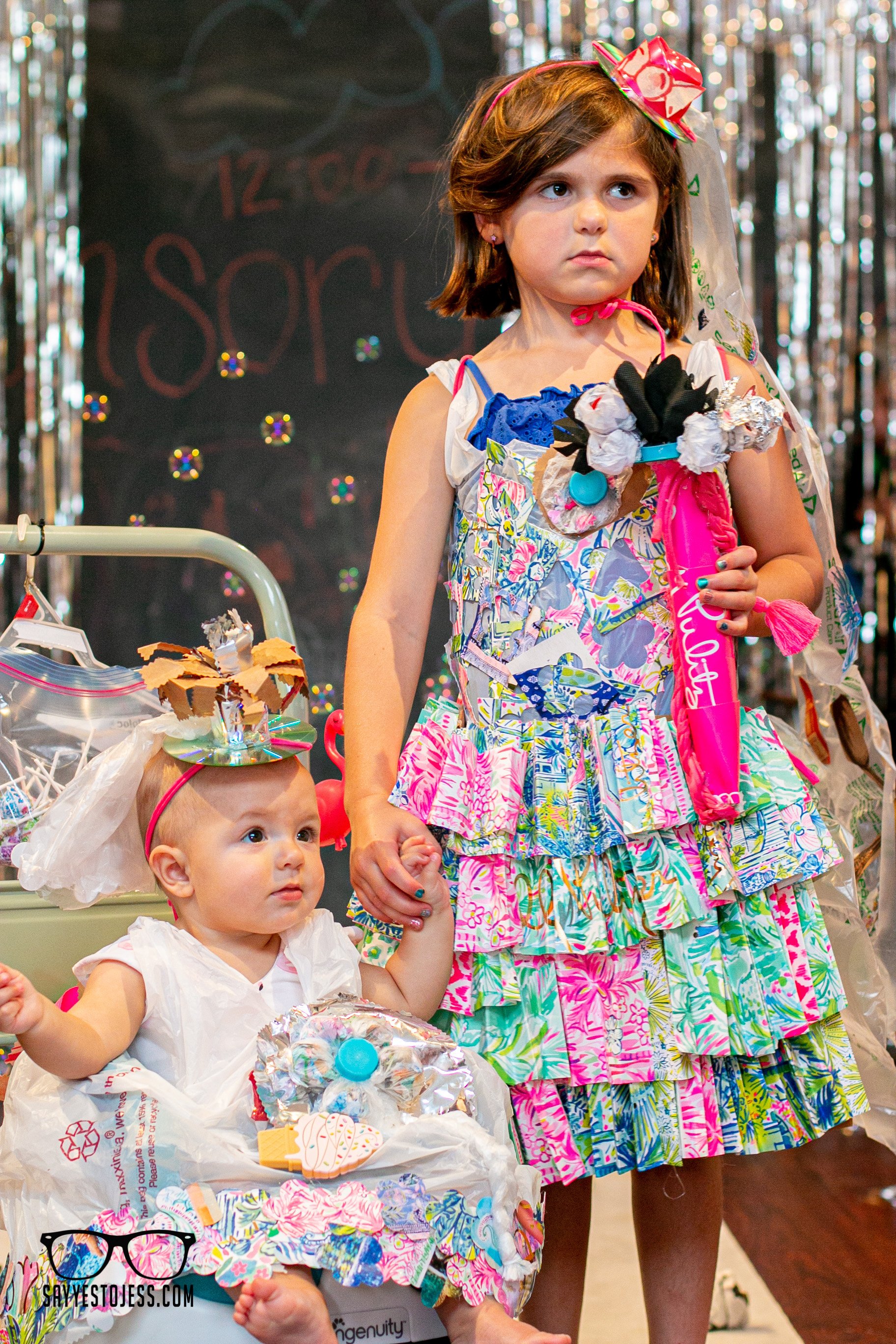 Two sisters in handmade recycled outfits holding hands on the runway with baby in a stroller at the Cincinnati trashion show