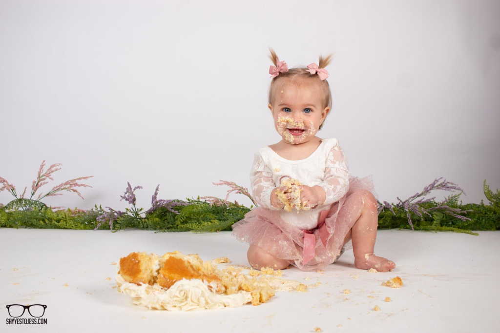 Toddler girl in a pink tutu with frosting on her face after smashing her 1st birthday cake at Say Yes to Jess