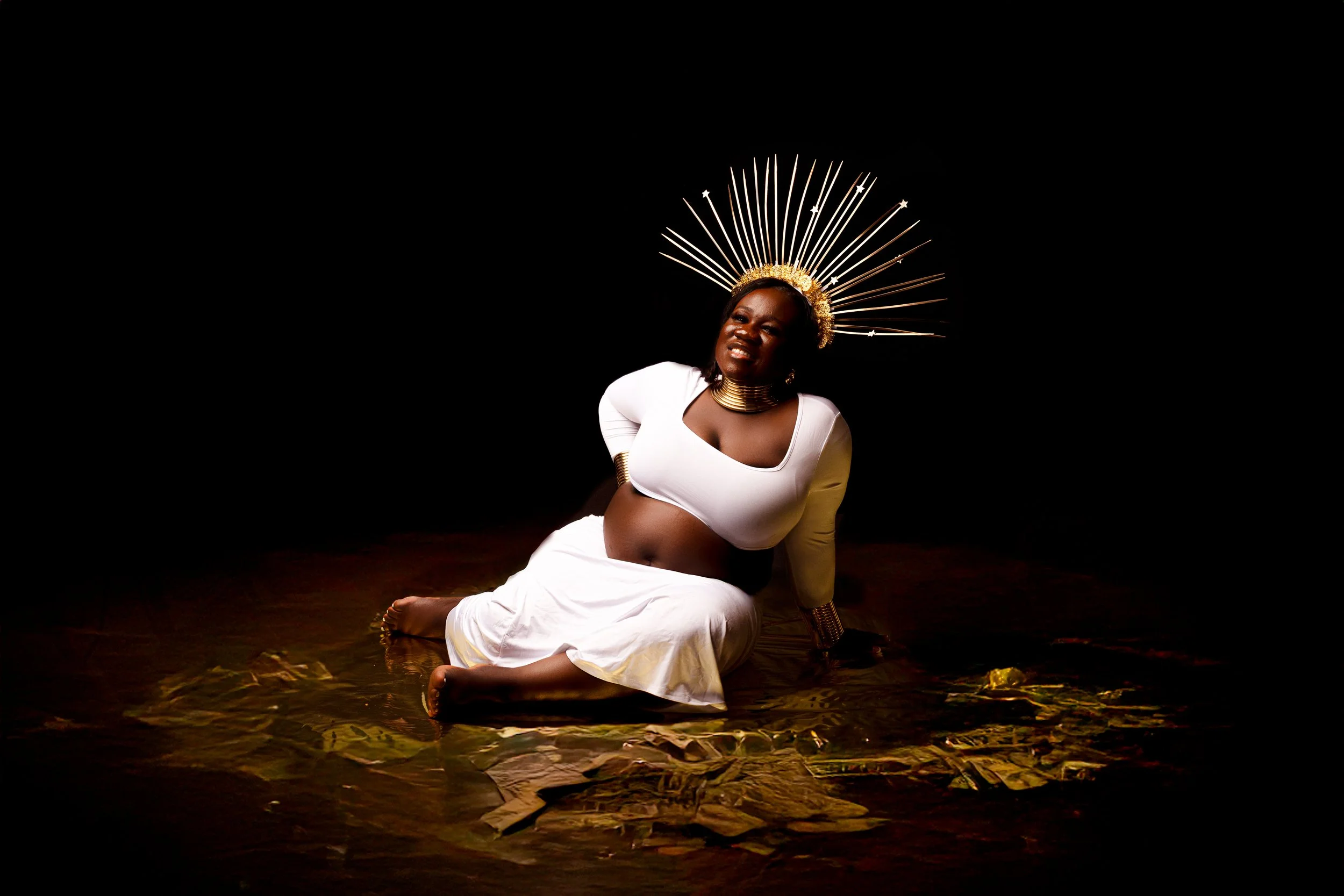 Creative maternity photoshoot with pregnant woman wearing a gold sunburst crown and gold accessories on a dramatic dark set with warm lighting in Cincinnati studio