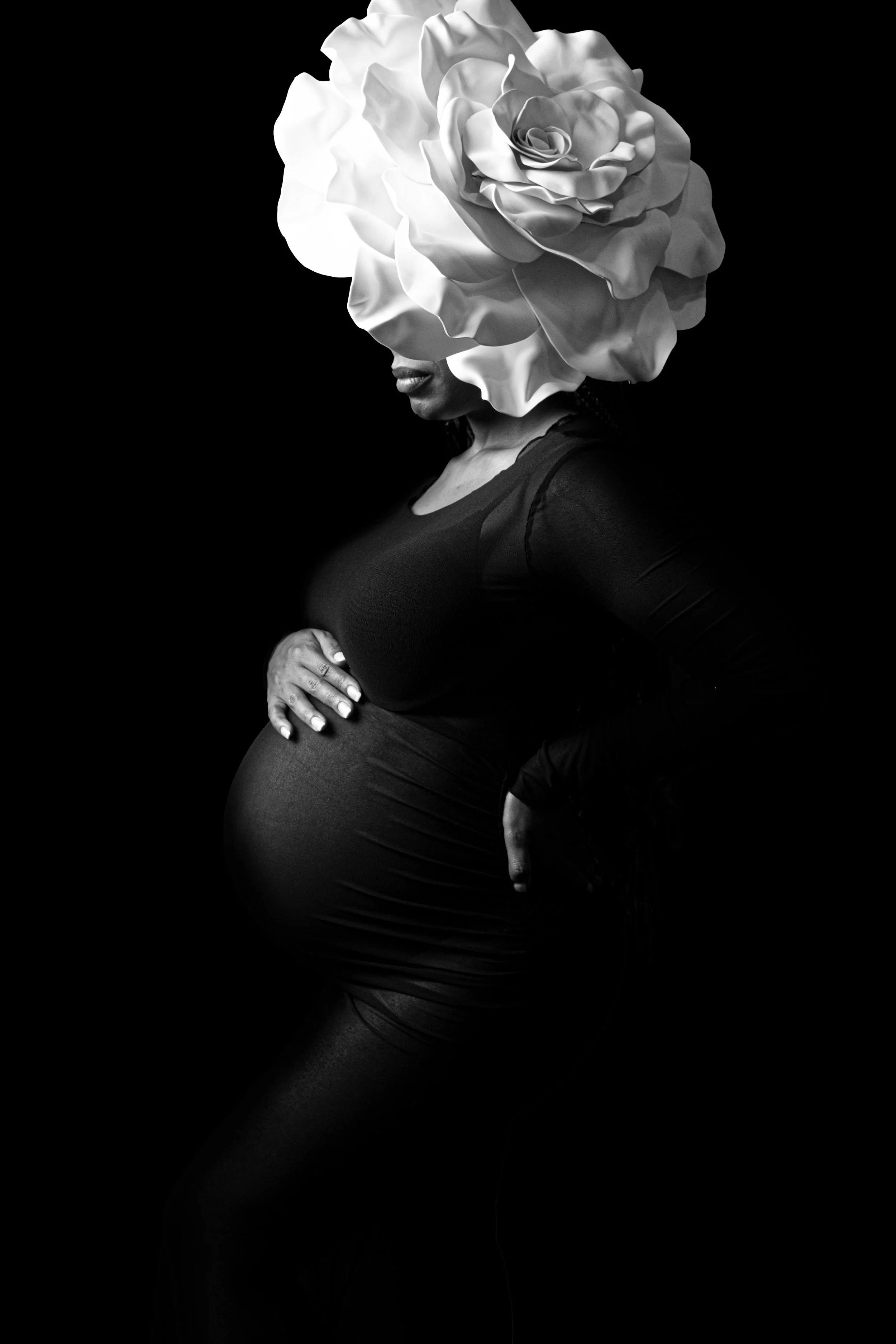 Pregnate woman wearing a giant floral hat that covers a portion of her face. The photograph is taken as a partial sillhouett on black background.