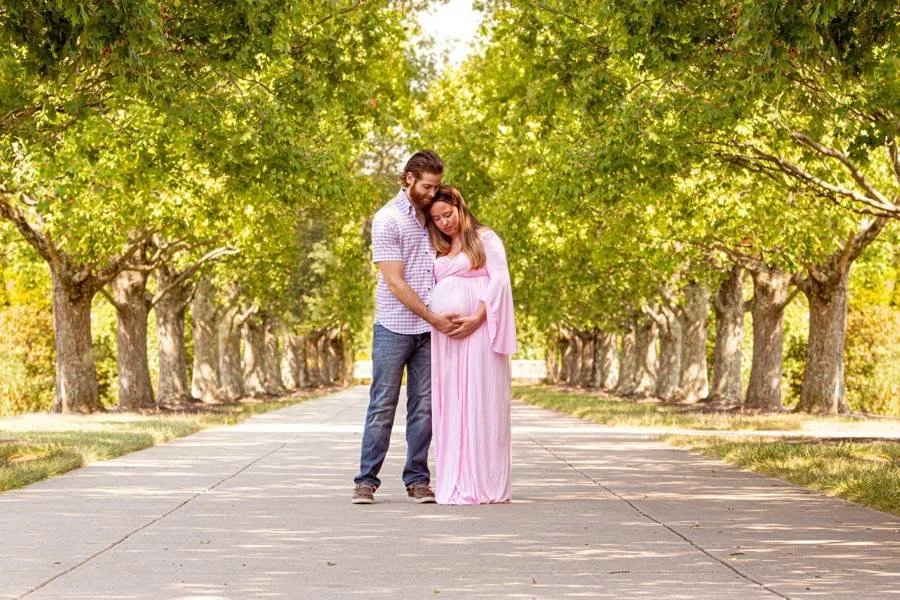 A couple standing along a shady tree line at ault park. They are expecting their first child and snuggled closely together for a cute shot.