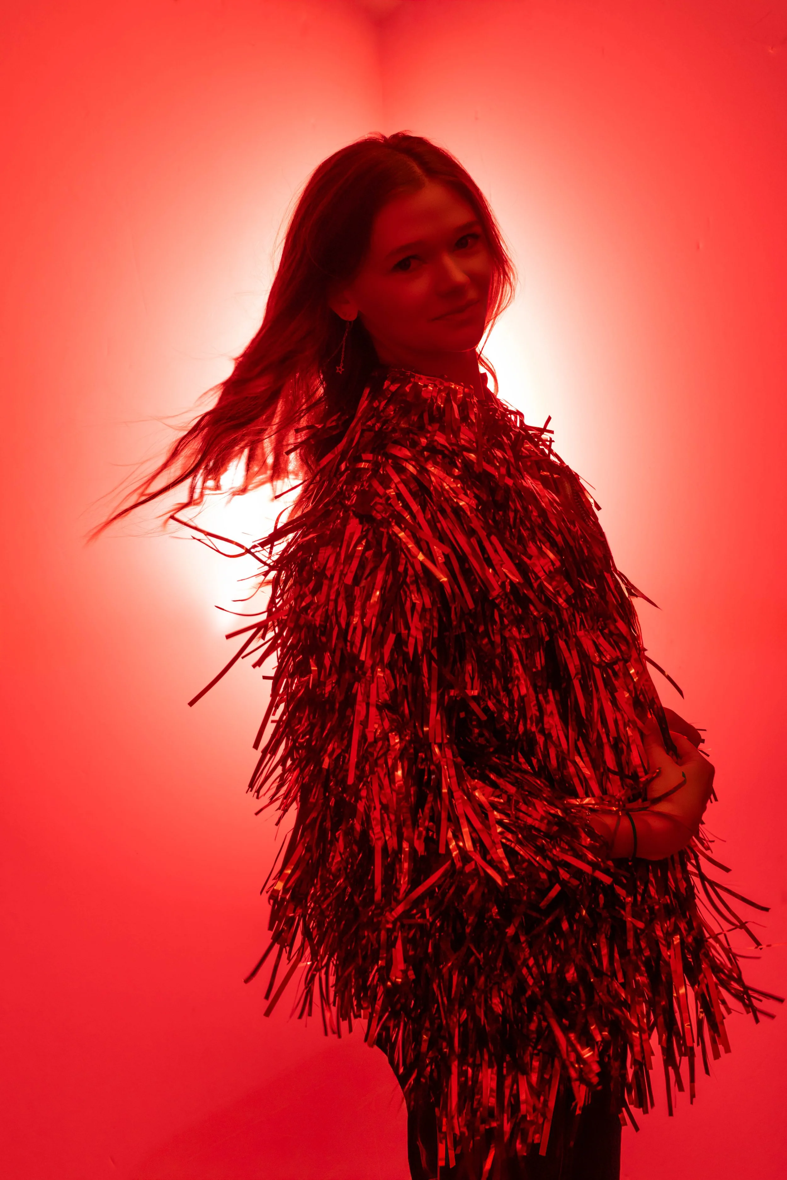 A red colored image with tons of light exploding around the photo. As well as a young girl turing towards the  camera in a shaggy glittering jacket. The jacket is showing movement as she turns.