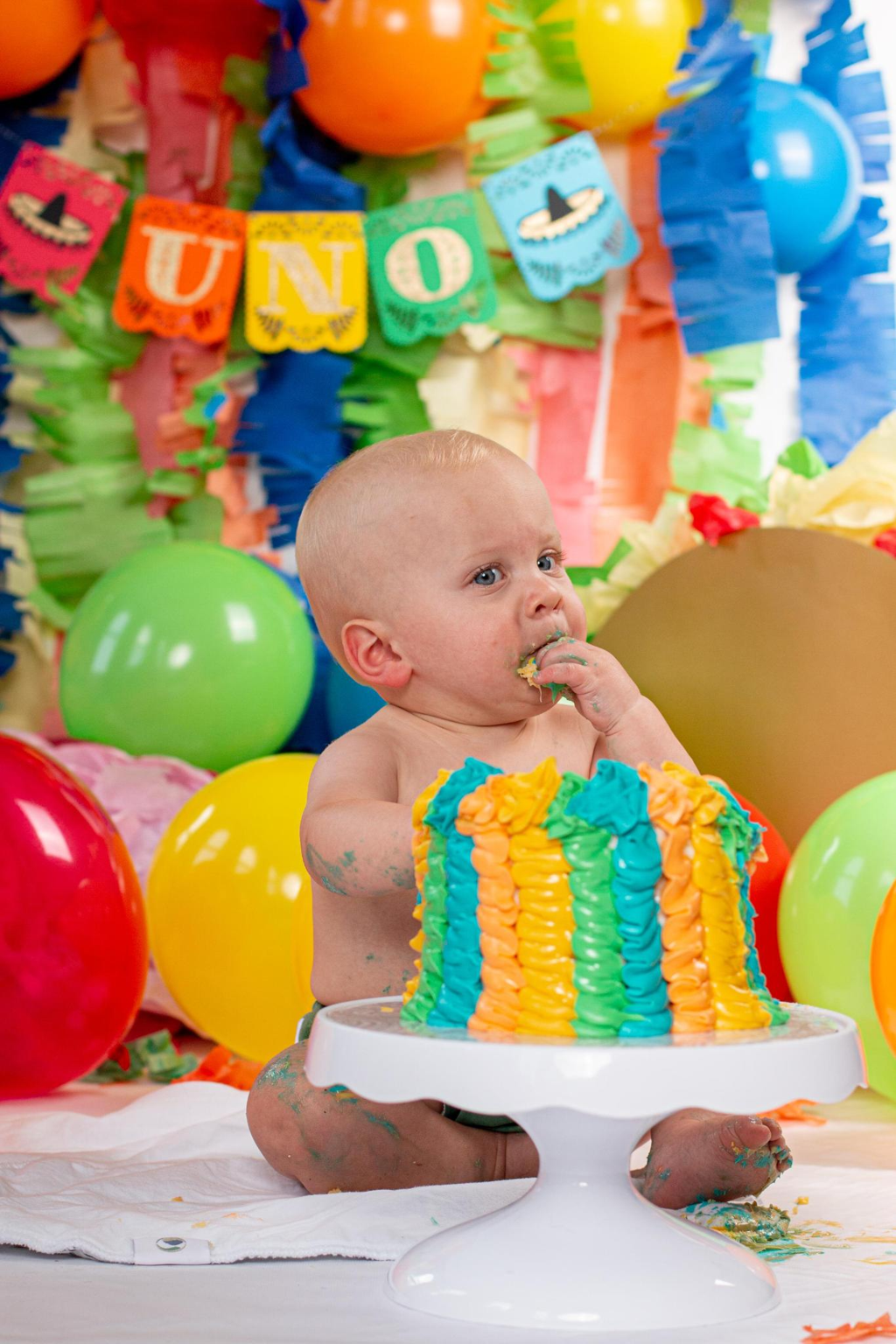 Baby at a fiesta themed 1st birthday cake smash with a rainbow cake and colorful balloon backdrop