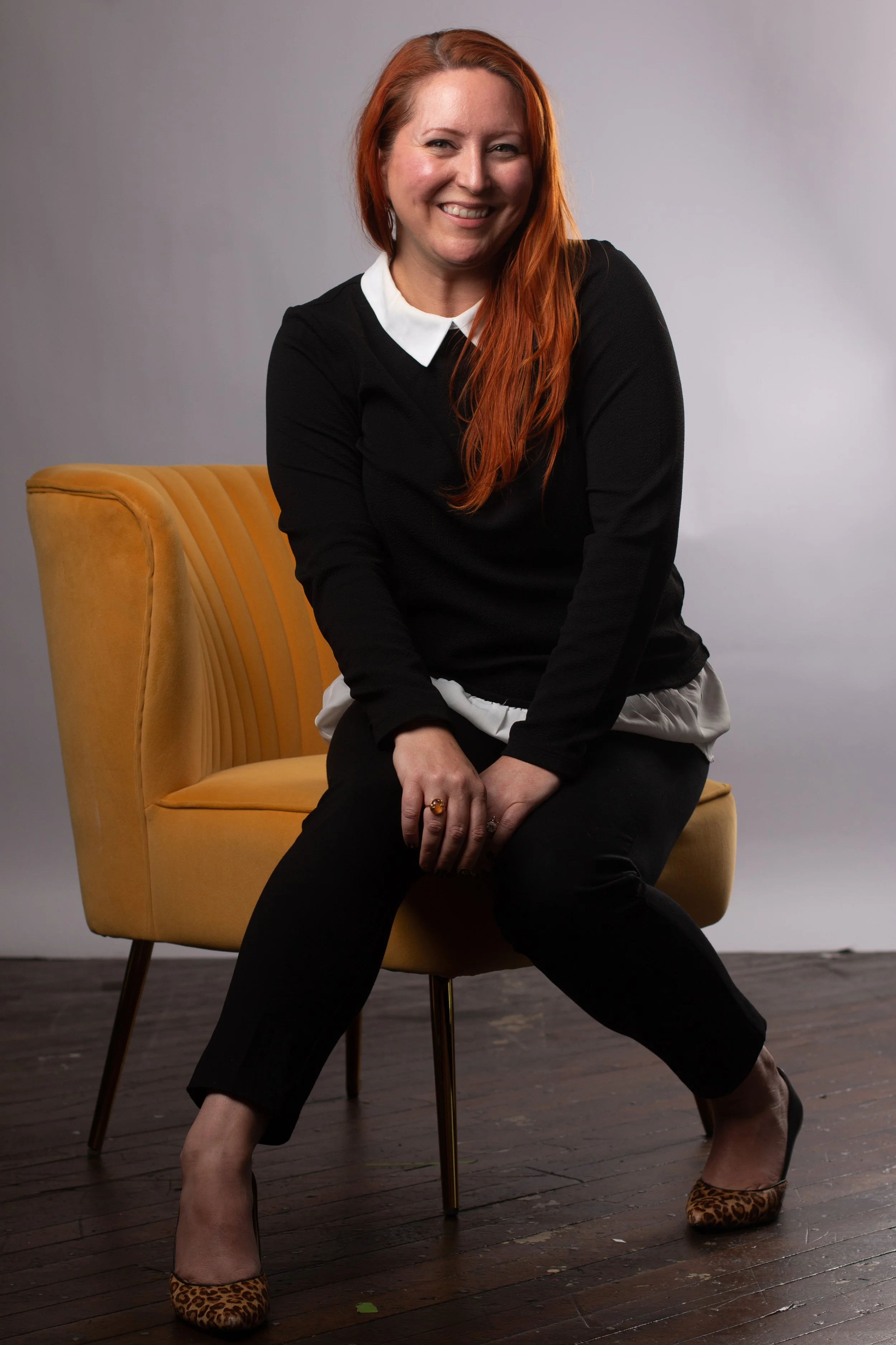 Professional headshot of a woman with red hair sitting on a mustard yellow chair smiling at the camera at Say Yes to Jess Cincinnati studio
