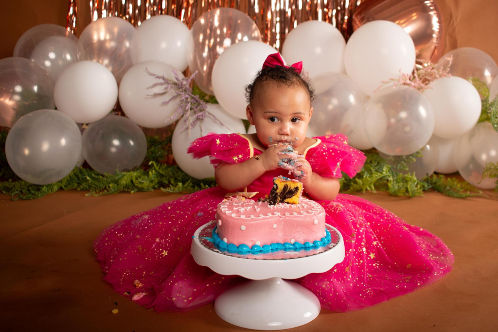 Baby girl in a hot pink dress at a 1st birthday cake smash with a white balloon arch and rose gold backdrop in Cincinnati