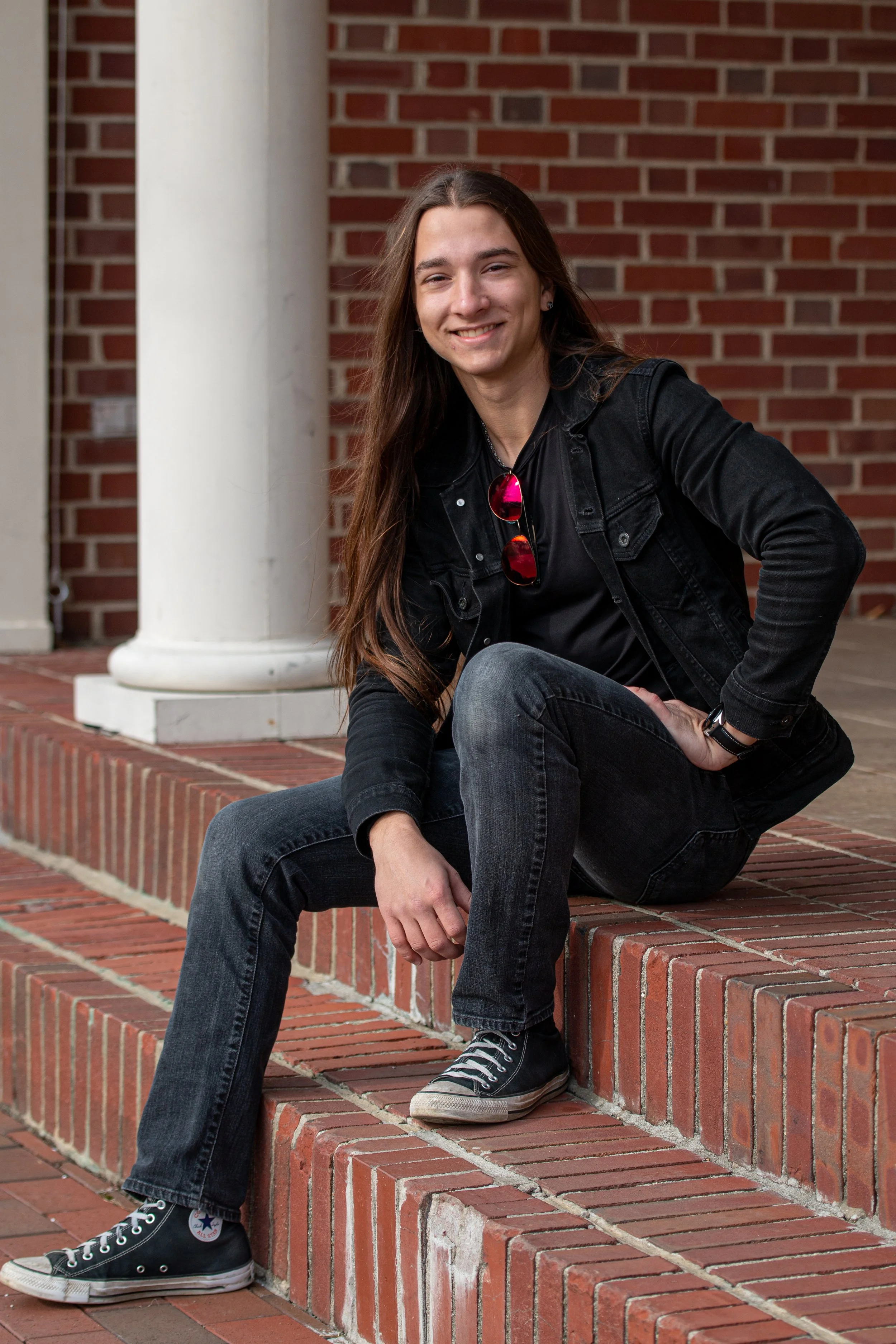 Senior pictures of guy with long hair in black jacket sitting on brick steps in relaxed pose