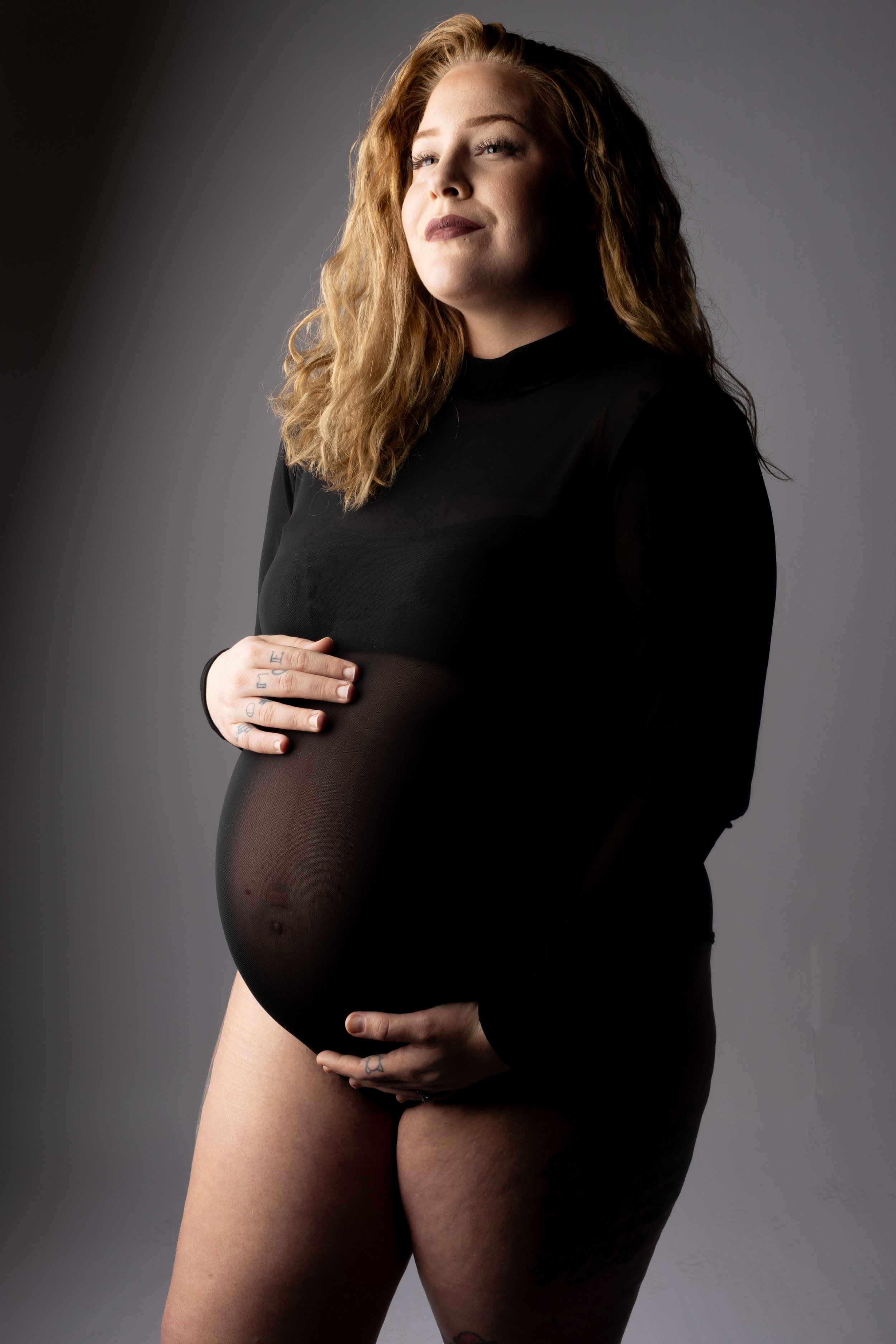 Bold maternity portrait of confident pregnant woman in sheer black top cradling her bump with moody studio lighting by Cincinnati maternity photographer Say Yes to Jess