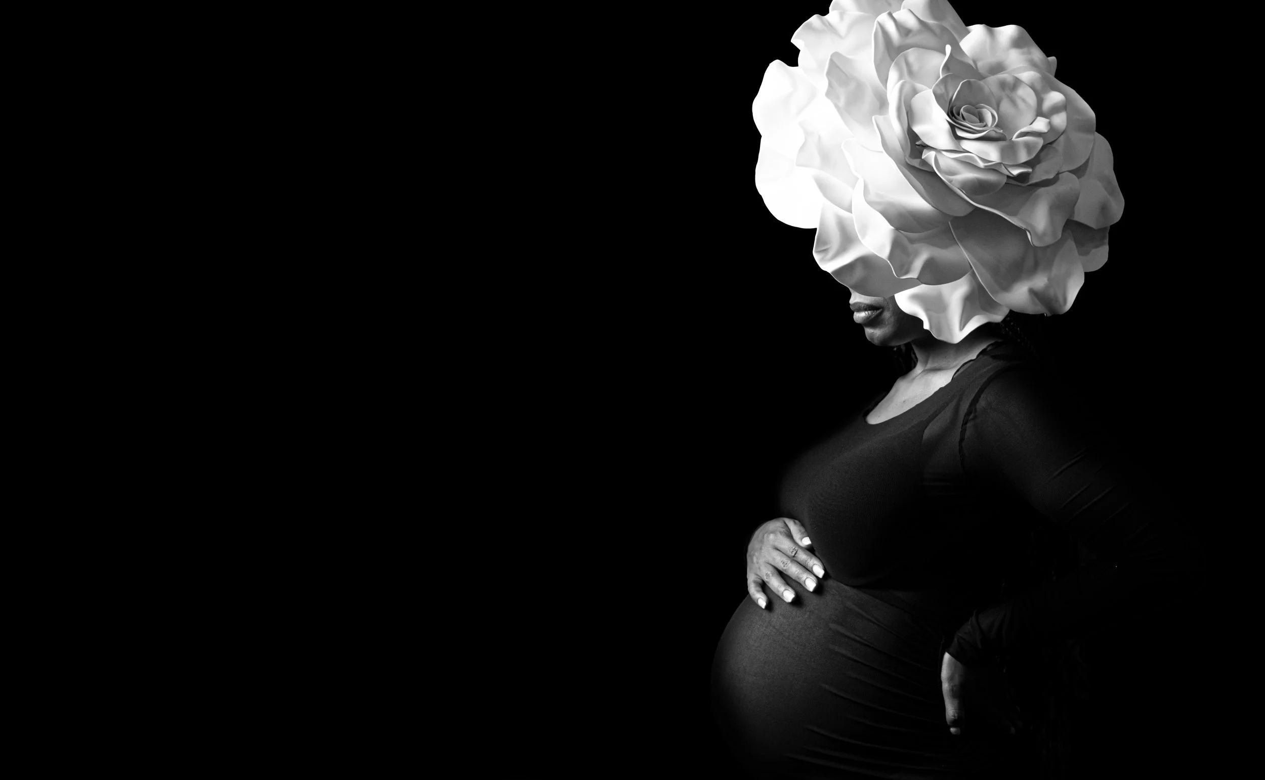 Artistic black and white maternity photo of pregnant woman wearing an oversized flower headpiece cradling her bump against a dark background by Say Yes to Jess Cincinnati photographer