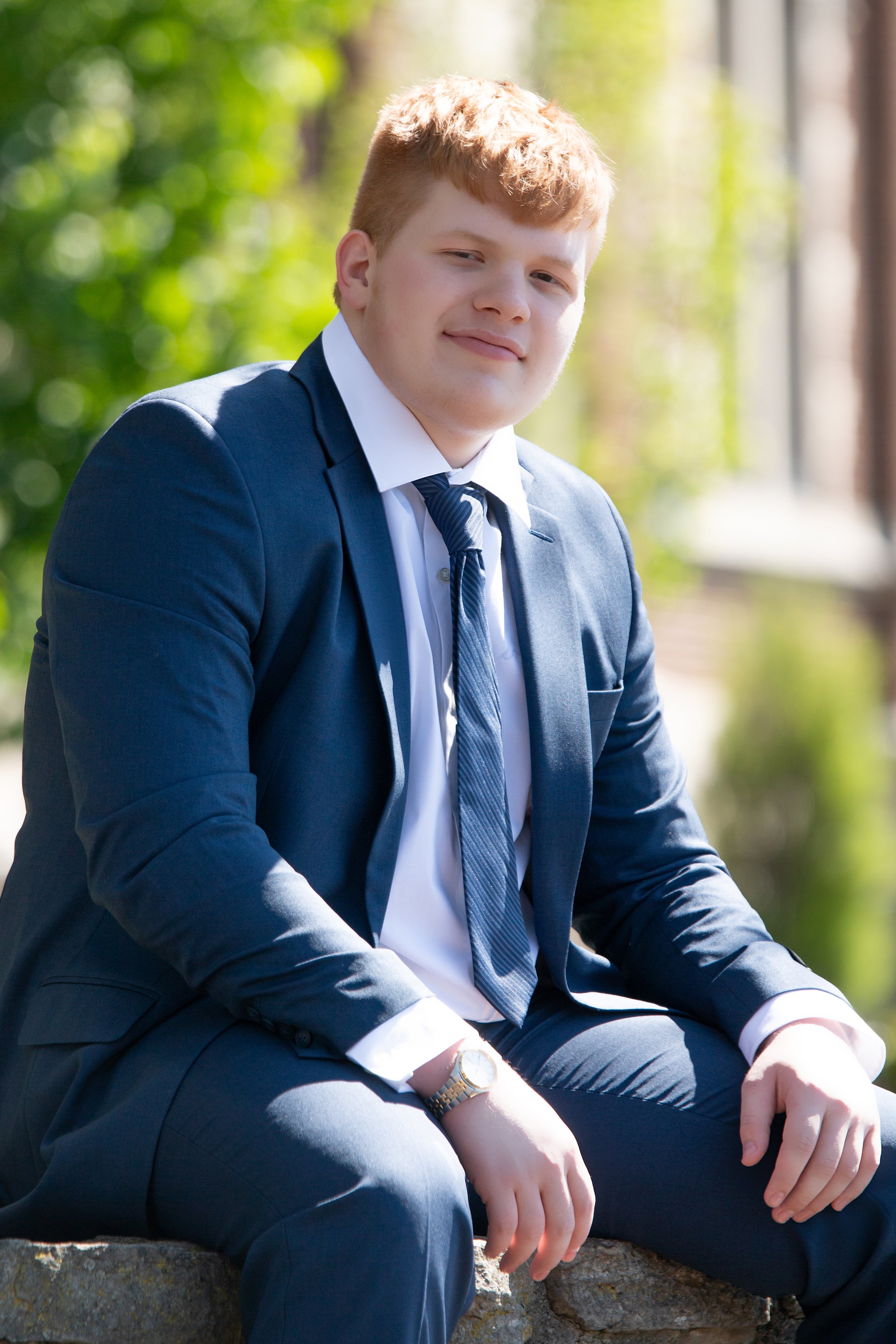 High school senior portrait of young man in navy blue suit and tie with outdoor greenery background