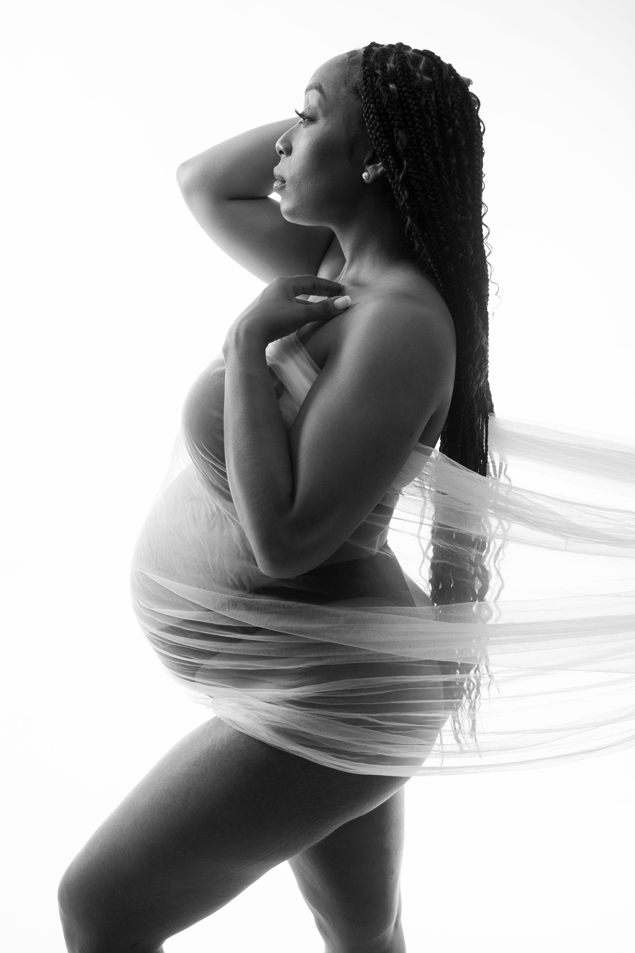 A stunning women of color standing with sheer fabric pulled to one side of the image to showcase her baby belly. Image is in black and white. Photographed on a white background.
