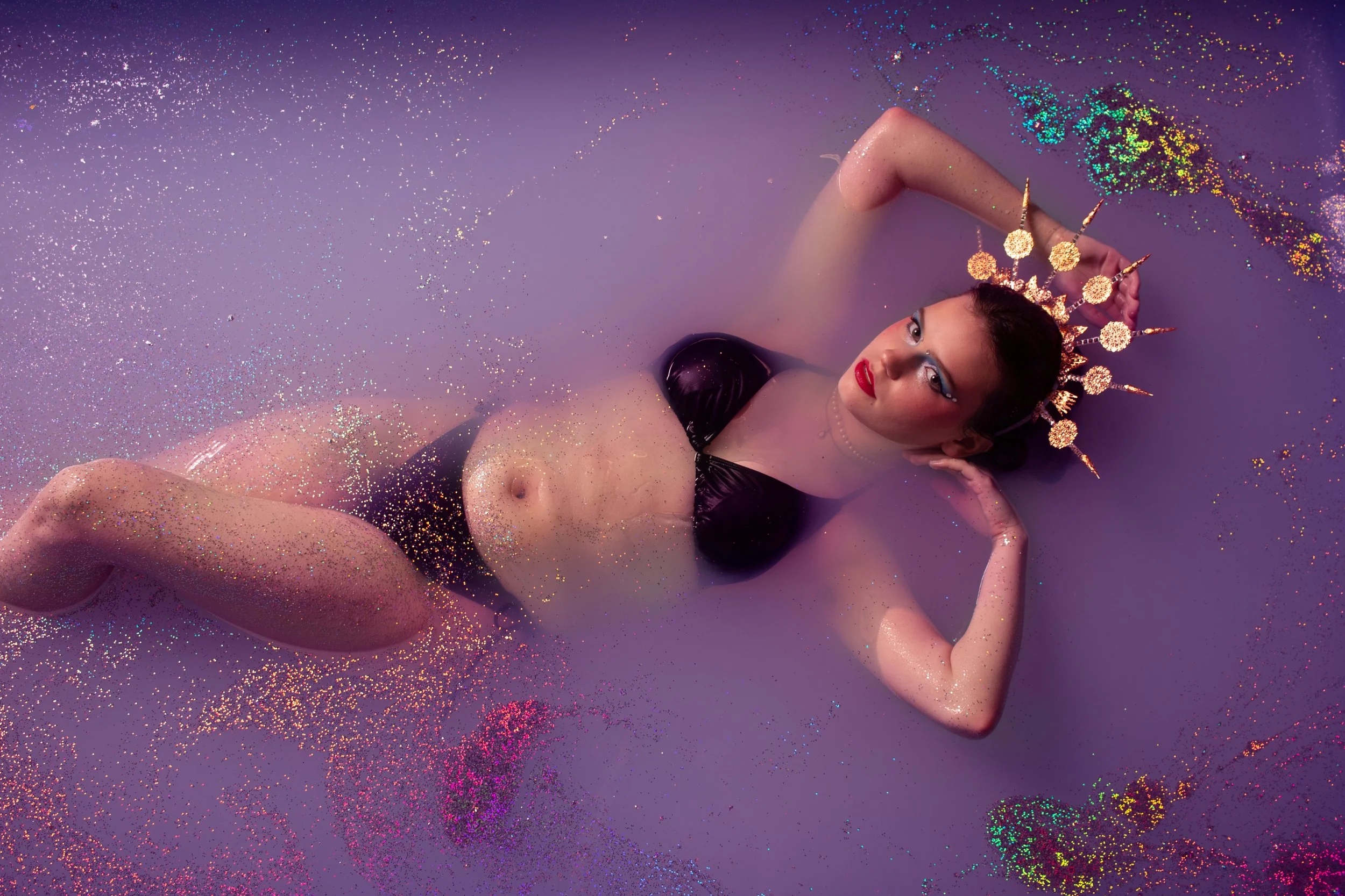 Woman in a black bikini with a gold floral crown lying in a lavender glitter milk bath during a floral milk bath portrait session at Say Yes to Jess Cincinnati