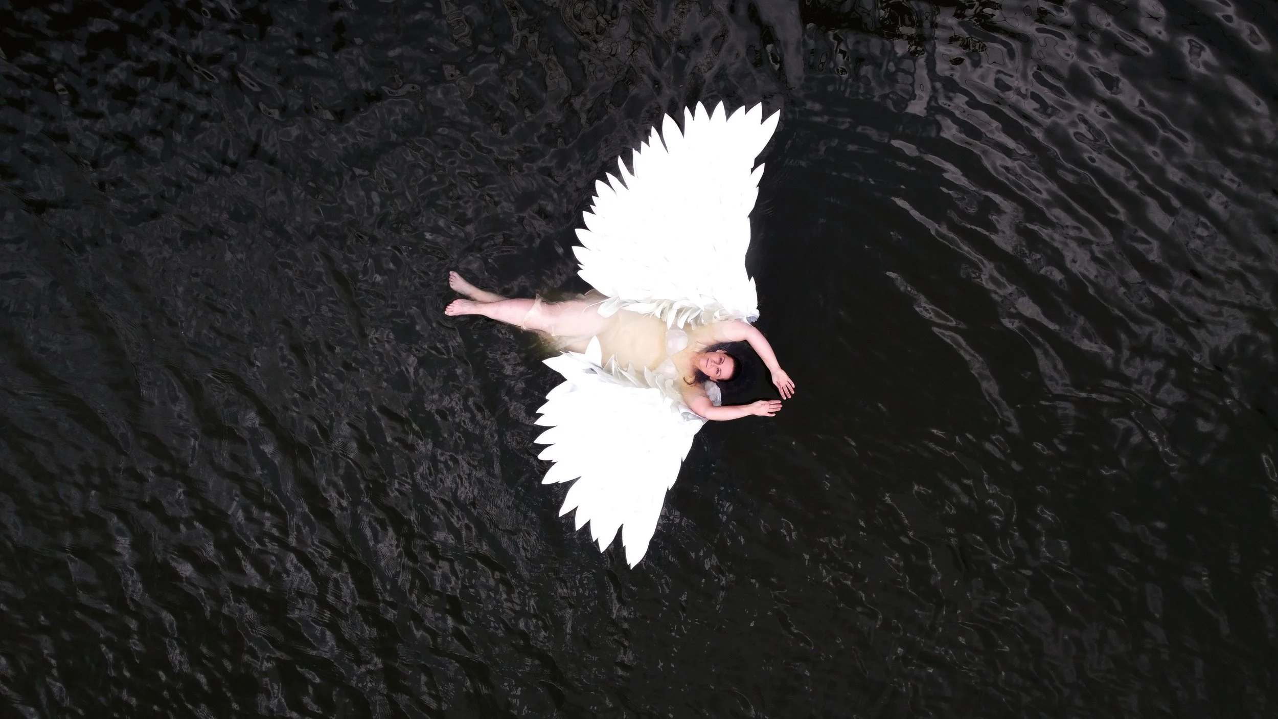 Woman lying on dark water with large white feathered angel wings spread out around her during a Celebrate HER empowerment session at Say Yes to Jess Cincinnati