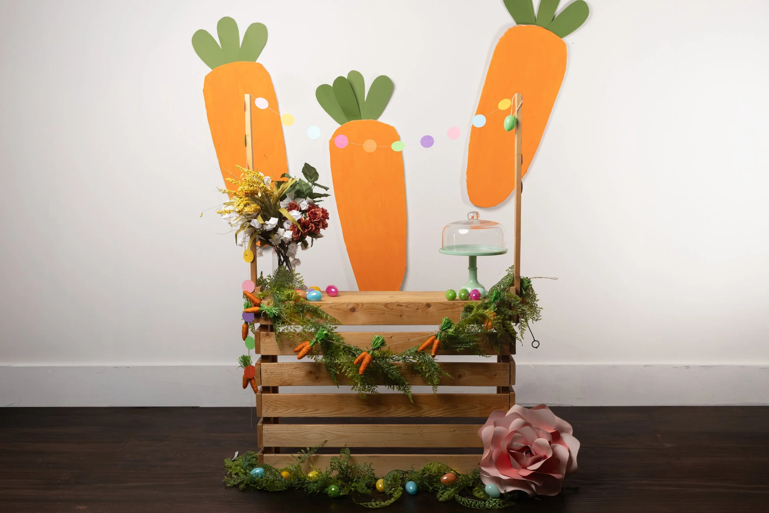 Custom hand-built Easter photography set featuring a wooden crate display with giant hand-painted orange carrots on the wall, a mini carrot garland, pastel polka dot string lights, colorful Easter eggs, spring flowers, greenery, a mint cake stand.
