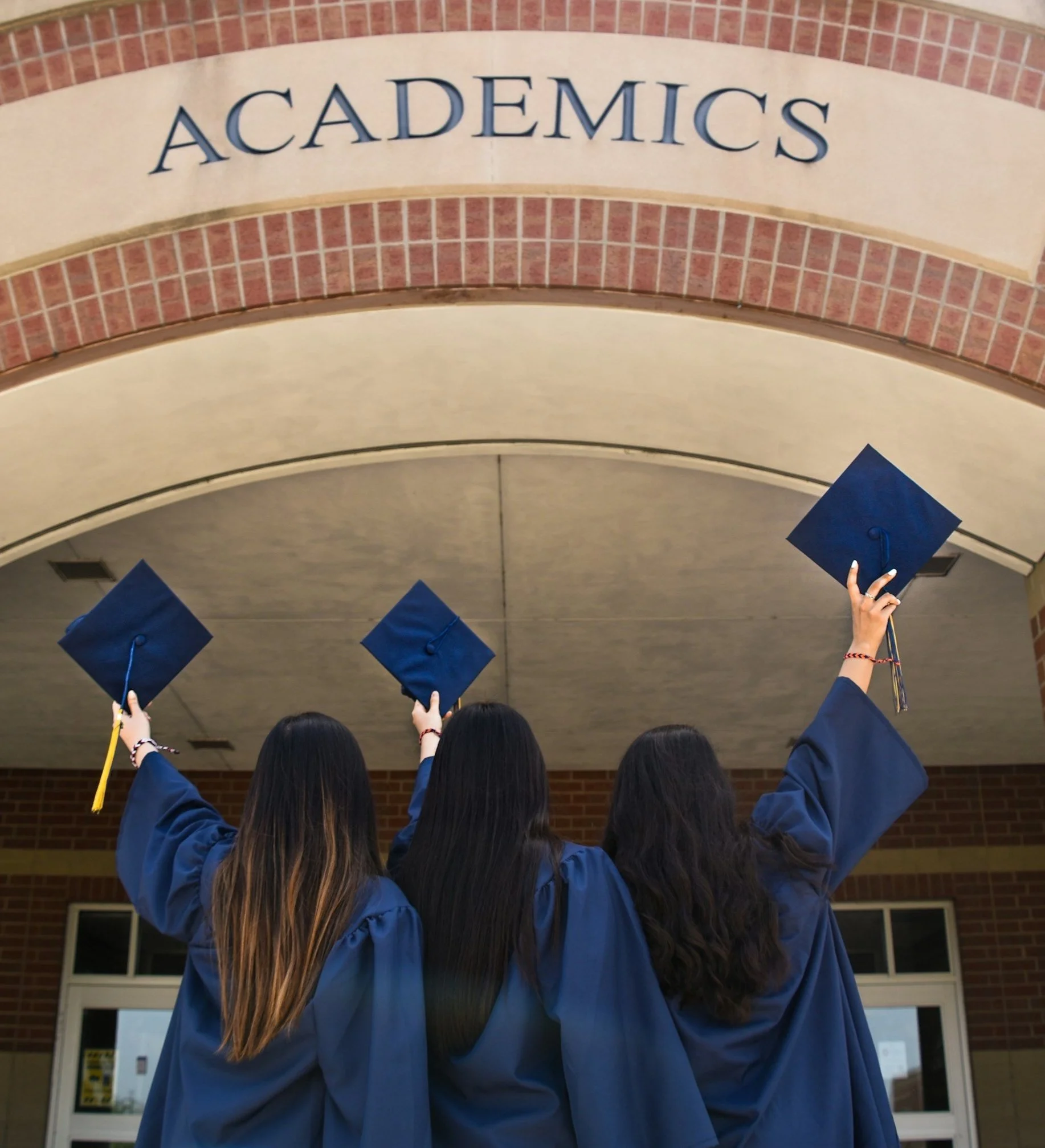 Three graduates in blue gowns holding caps under 'Academics' arch.