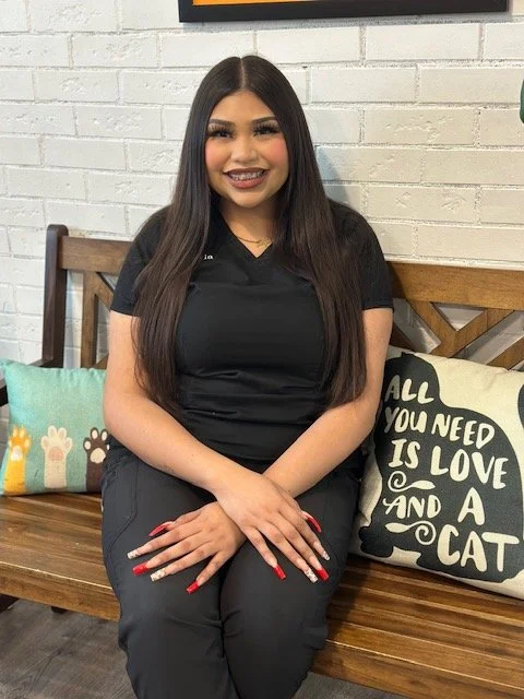 A woman with long dark hair and braces sitting on a wooden bench with decorative animal pillows, wearing black scrubs and sneakers, against a white brick wall.