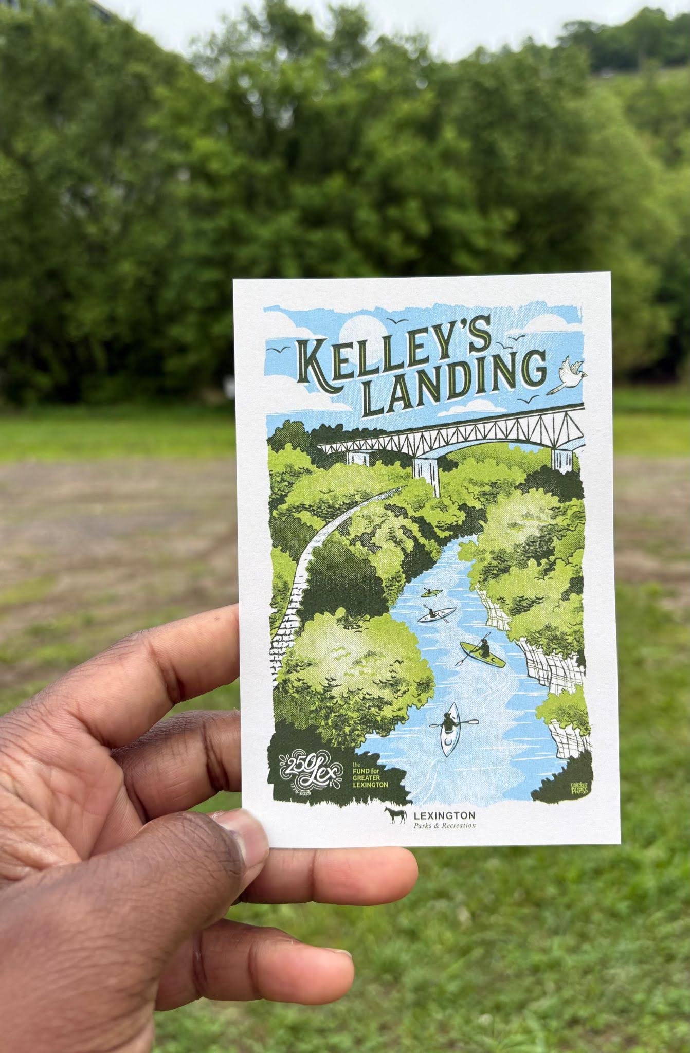 Hand holding an illustrated Kelley’s Landing informational postcard against a blurred outdoor park background.