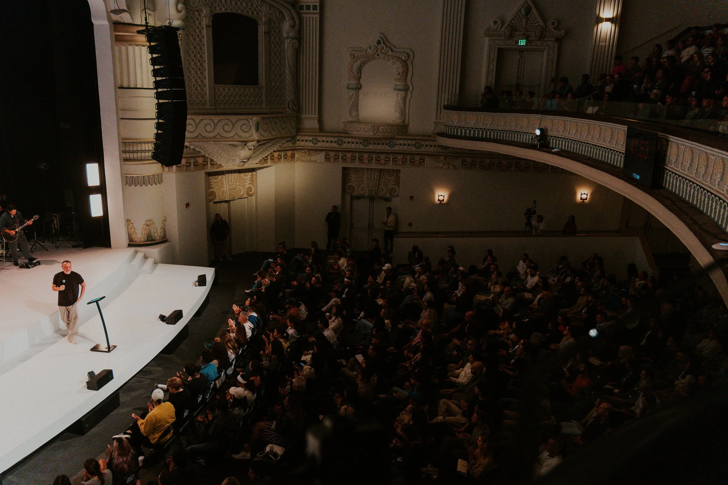 A speaker stands on a white stage in a large auditorium, addressing an audience seated in rows. The auditorium features ornate architectural details on the walls and curved upper balconies, with many people watching the presentation.