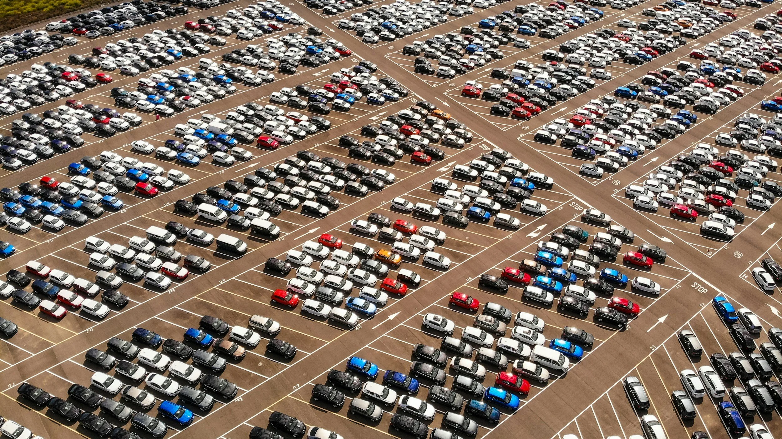 Aerial view of a large parking lot filled with numerous white, black, silver, red, blue, and other colored cars parked in organized rows.