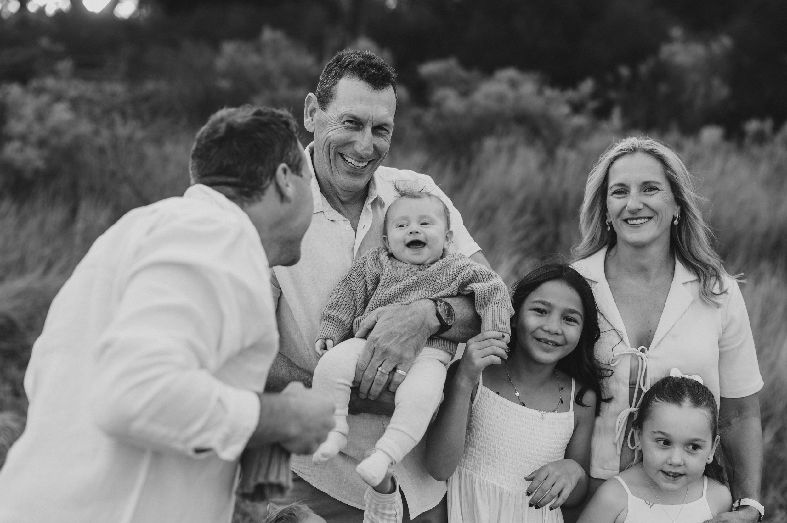 A black and white photo of a family group outdoors, with adults and children laughing and smiling. A man holds a baby while another person engages with the child. Two girls stand nearby, smiling, and there is a woman in the background. The scene is s