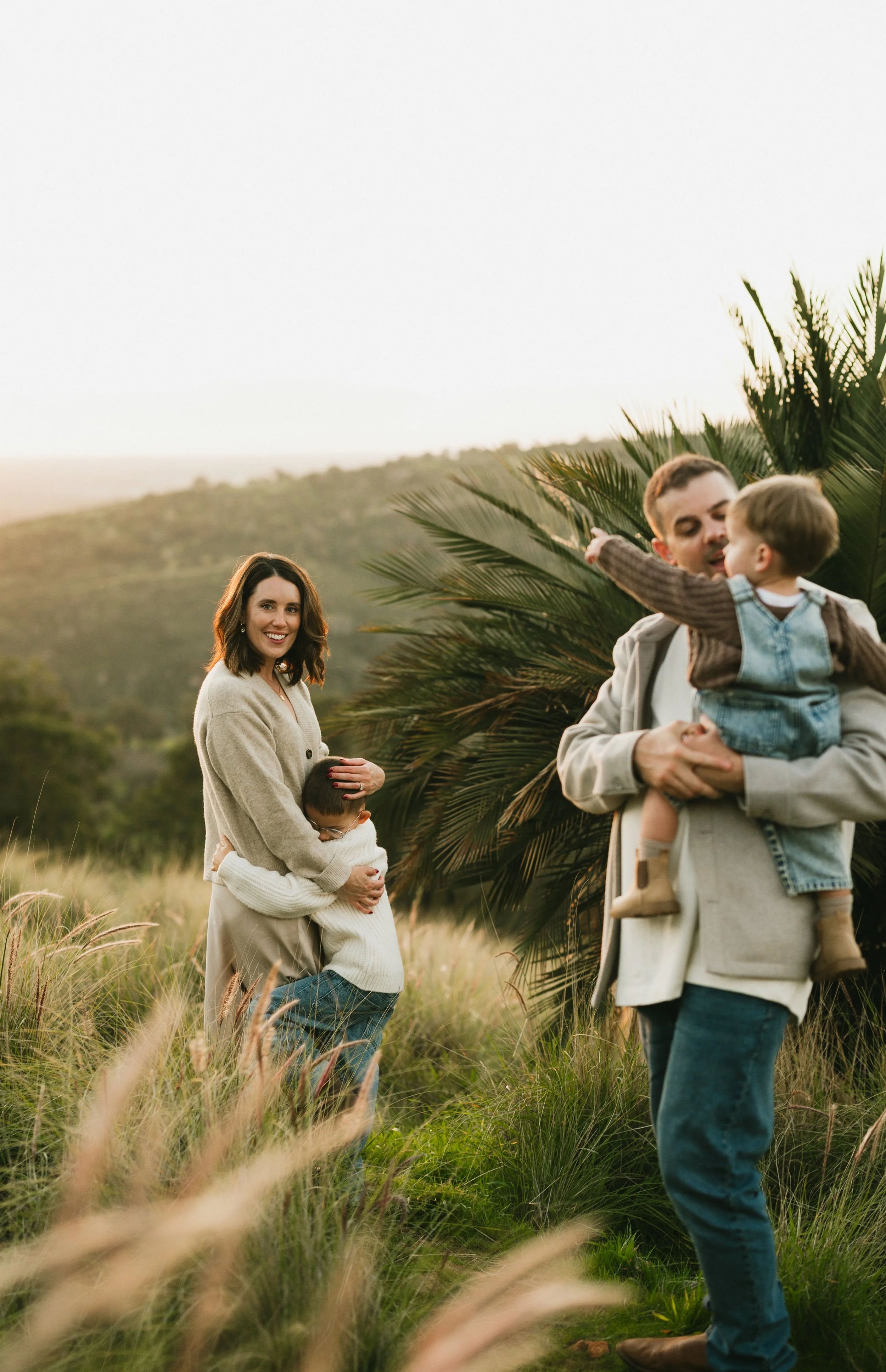 A family of three walking on a dirt road. The parents are holding hands with their young child, lifting them in the air. The background features trees and a sunny outdoor setting.
