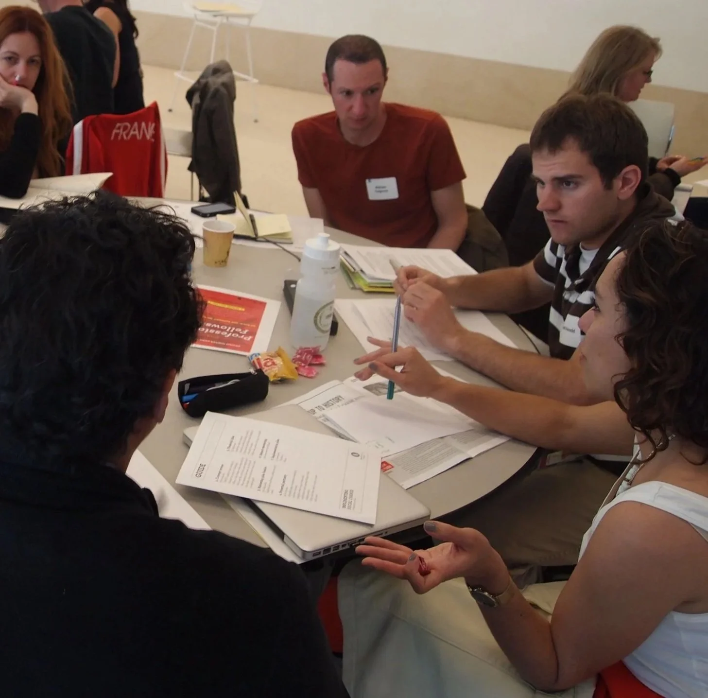 A group of people seated around a table during a meeting or workshop, with documents, notebooks, and drinks on the table.