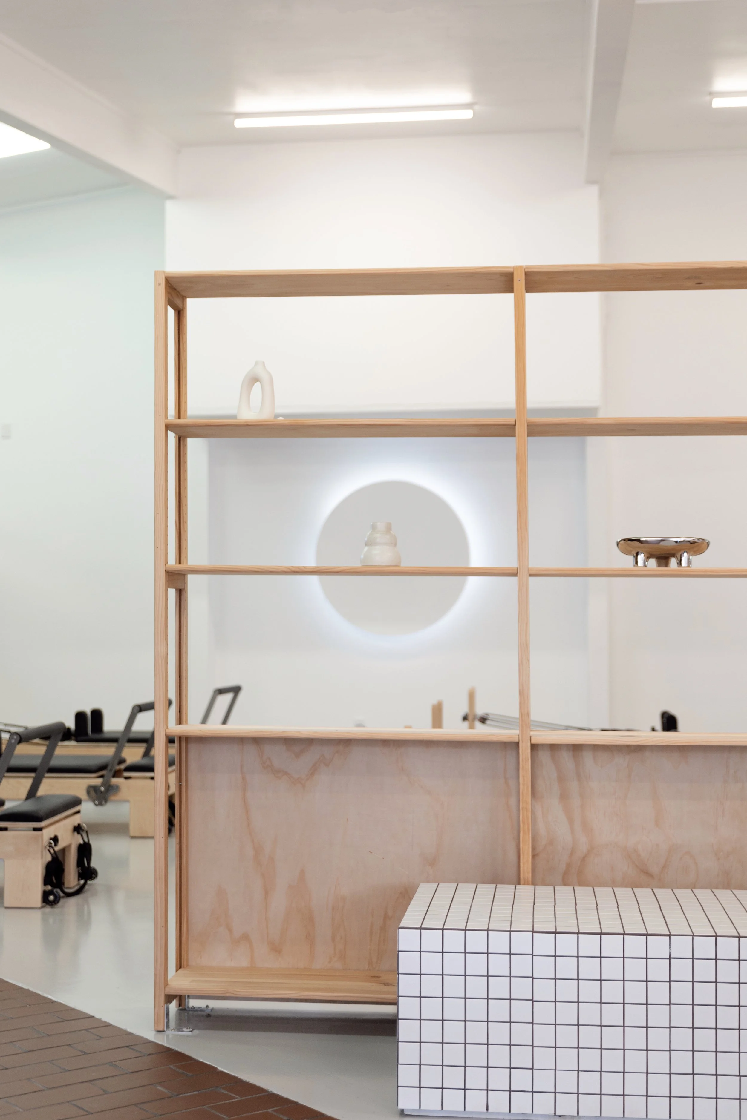 Core Club interior gallery space featuring a wooden open-frame shelving unit with objects. In the background, there is the Reformer Studio with a circular opening illuminated by a light.