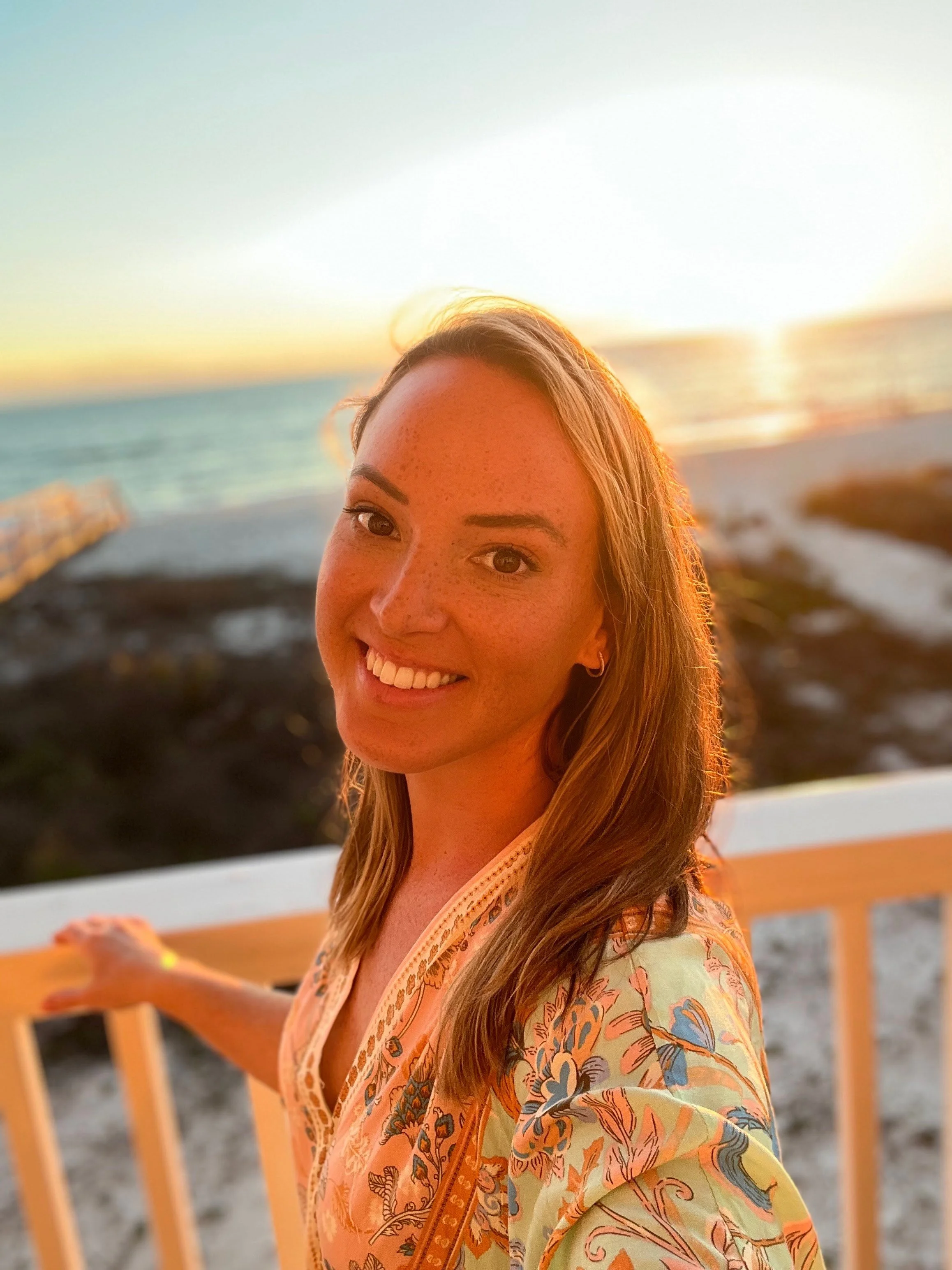 Woman smiling at the beach during sunset, wearing a floral top, with ocean and sky in the background.