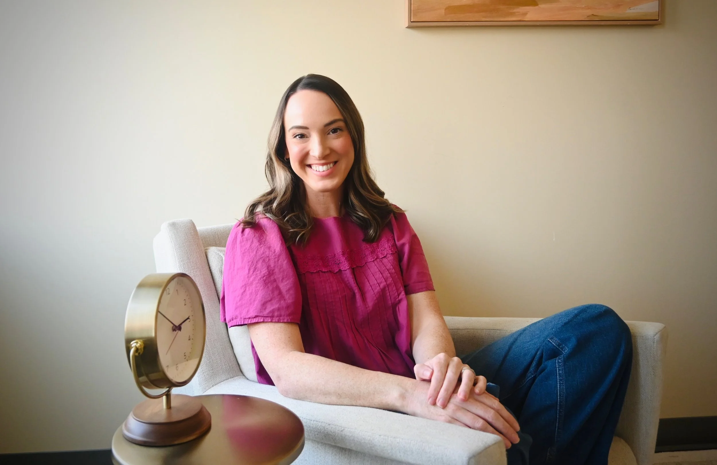 A young woman with shoulder-length brown hair, wearing a magenta blouse and blue jeans, smiling while sitting on a beige sofa. There is a round, gold-colored alarm clock on a wooden table in front of her and a plain, light-colored wall behind her.