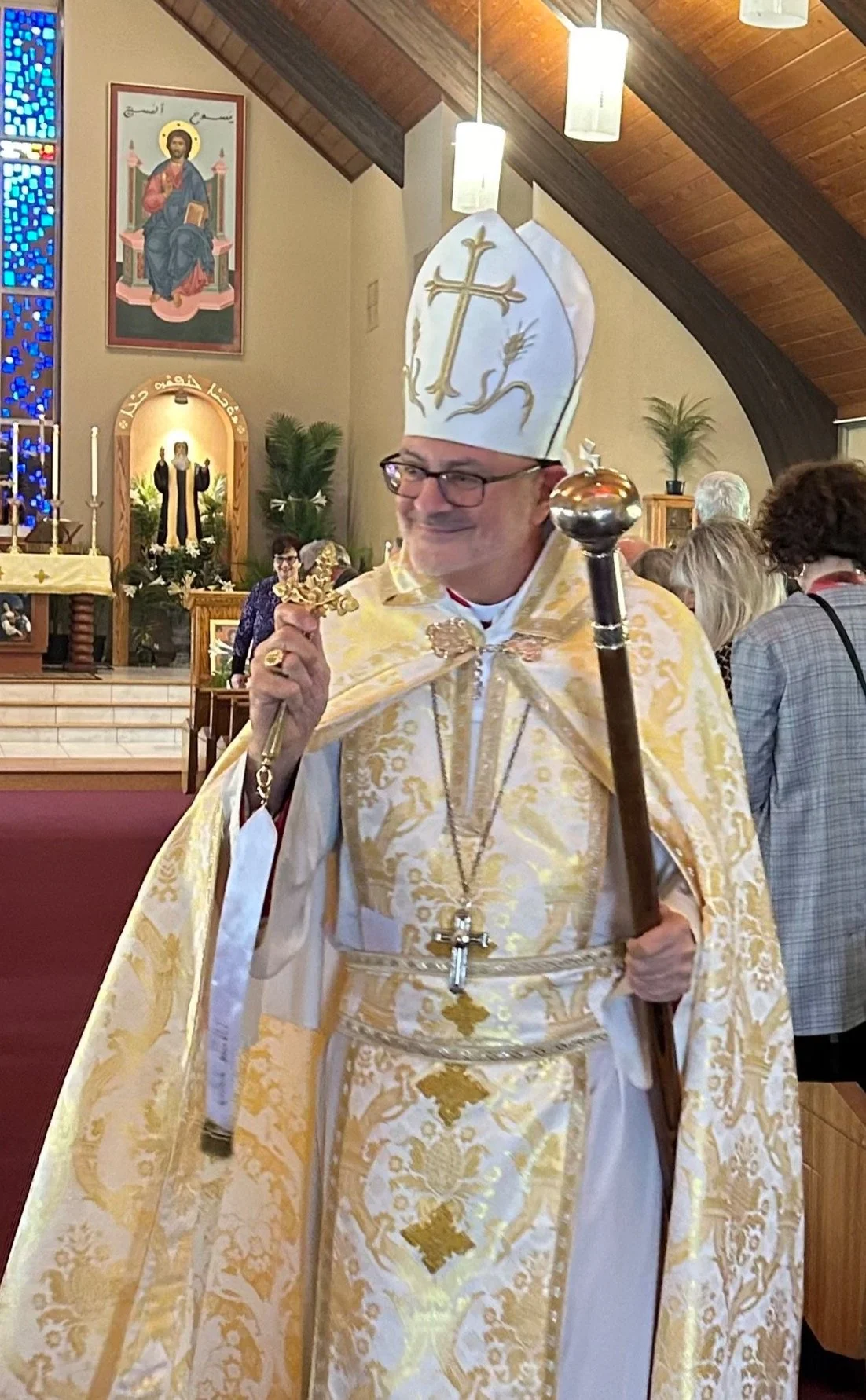 A bishop in ornate gold and white vestments, wearing a mitre decorated with a cross, holding a crosier and a gold cross, inside a church with people gathered for a service.
