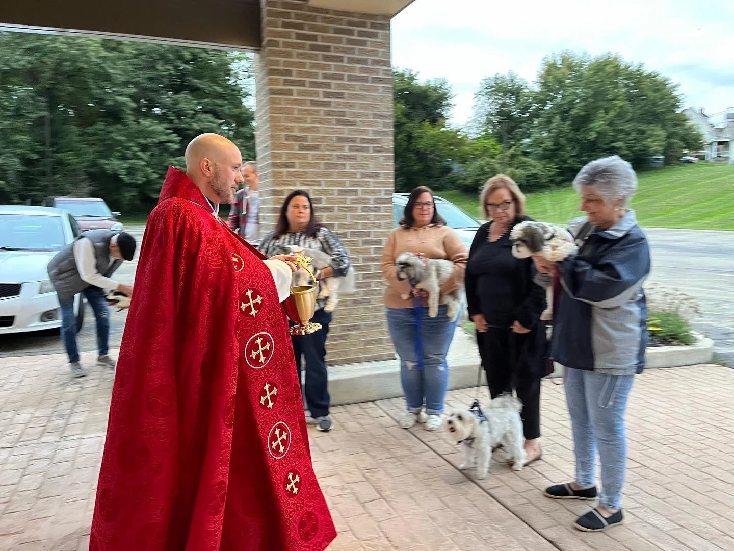 A priest in red robes holding a chalice and a cross, performing a blessing or ceremony with a group of women and their small dogs outside a building on a cloudy day.
