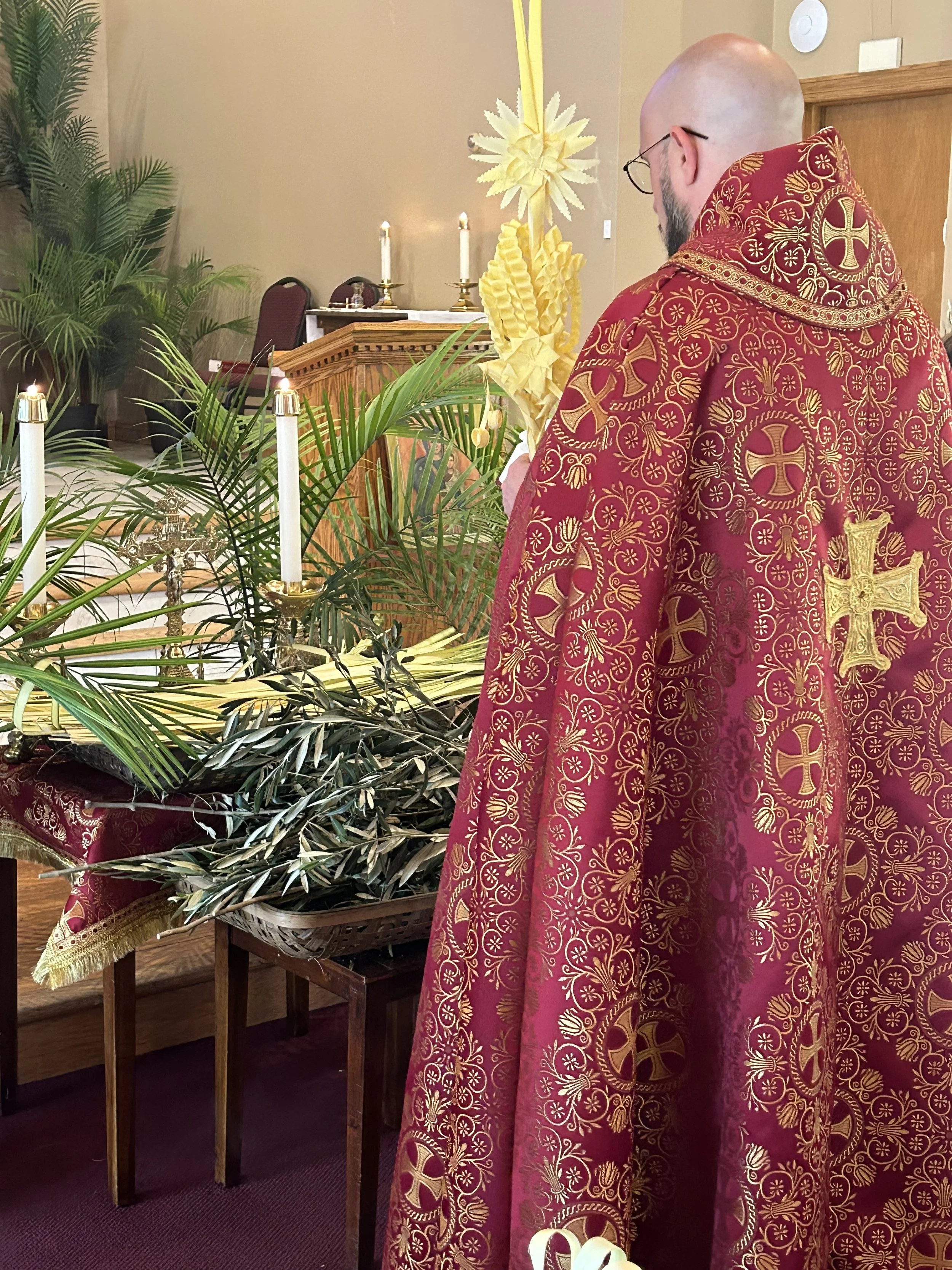 A priest wearing a red and gold embroidered vestment stands in front of an altar with lit candles, palm branches, and a flower arrangement, participating in a religious service.