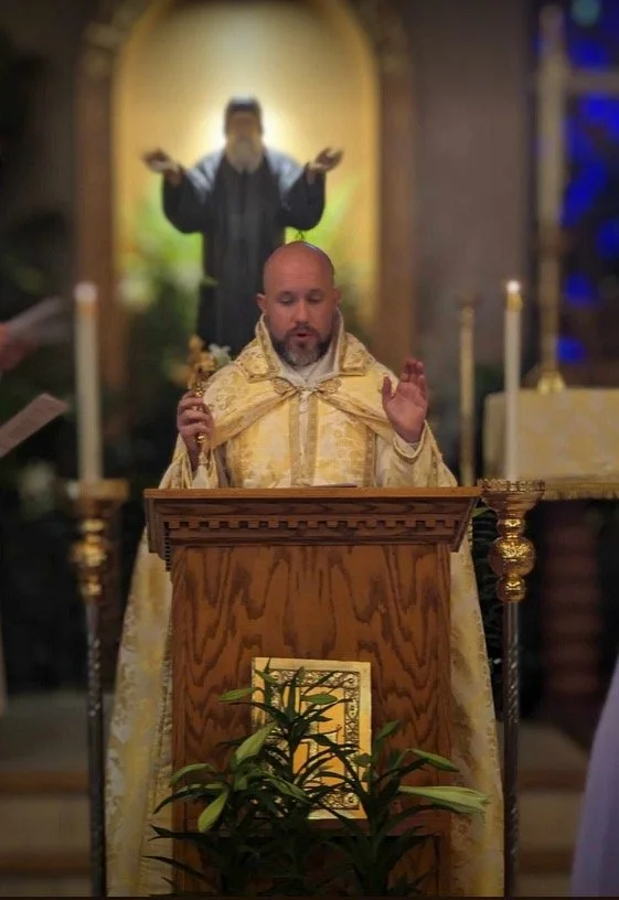 A priest in ornate gold vestments stands behind an altar with a wooden lectern, raising his right hand in prayer during a religious service, with a priest in black robes in the background with arms extended in a blessing gesture.