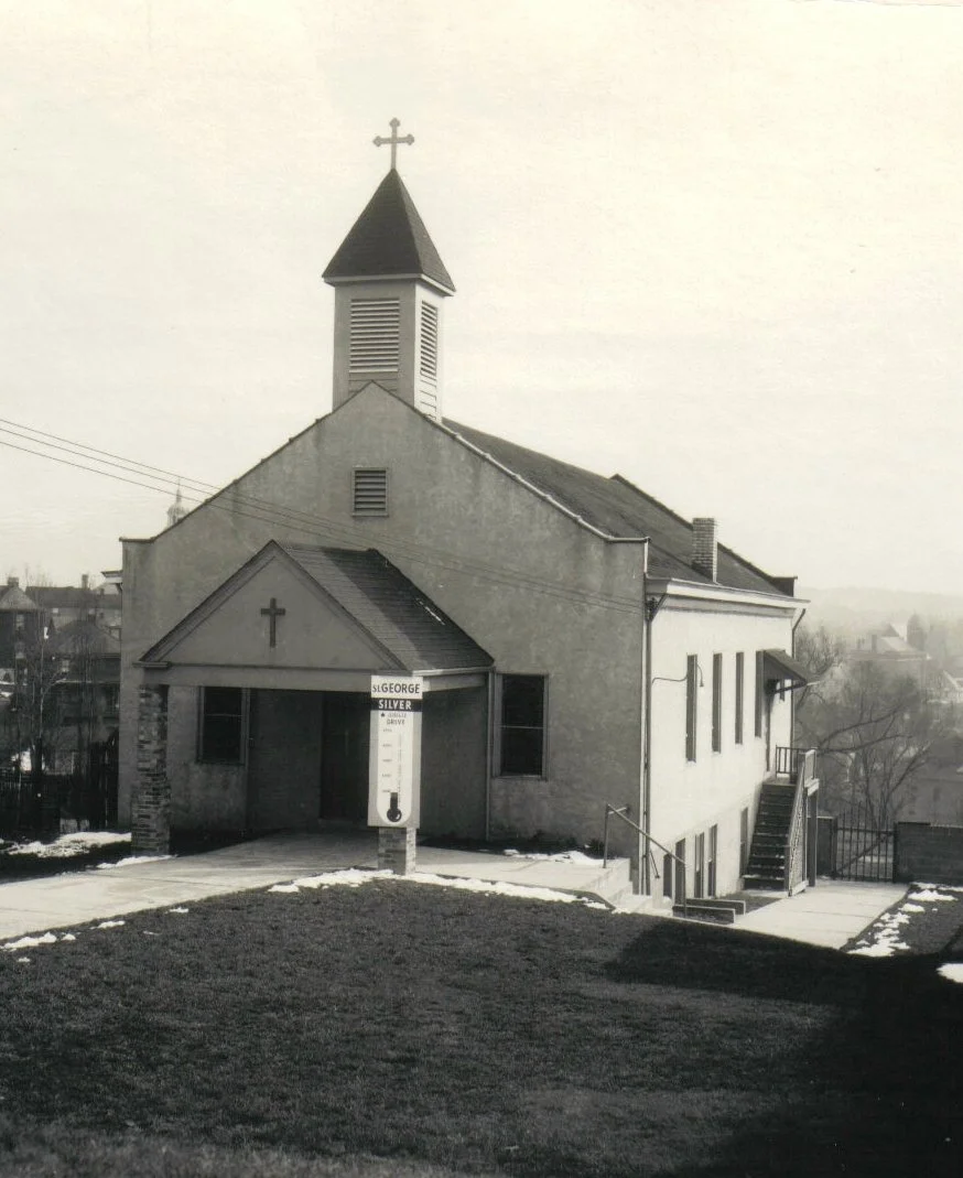 Black and white photo of a small church with a steeple and a cross on top, situated on a hill with patches of snow, and a sign for a gas station at the entrance.