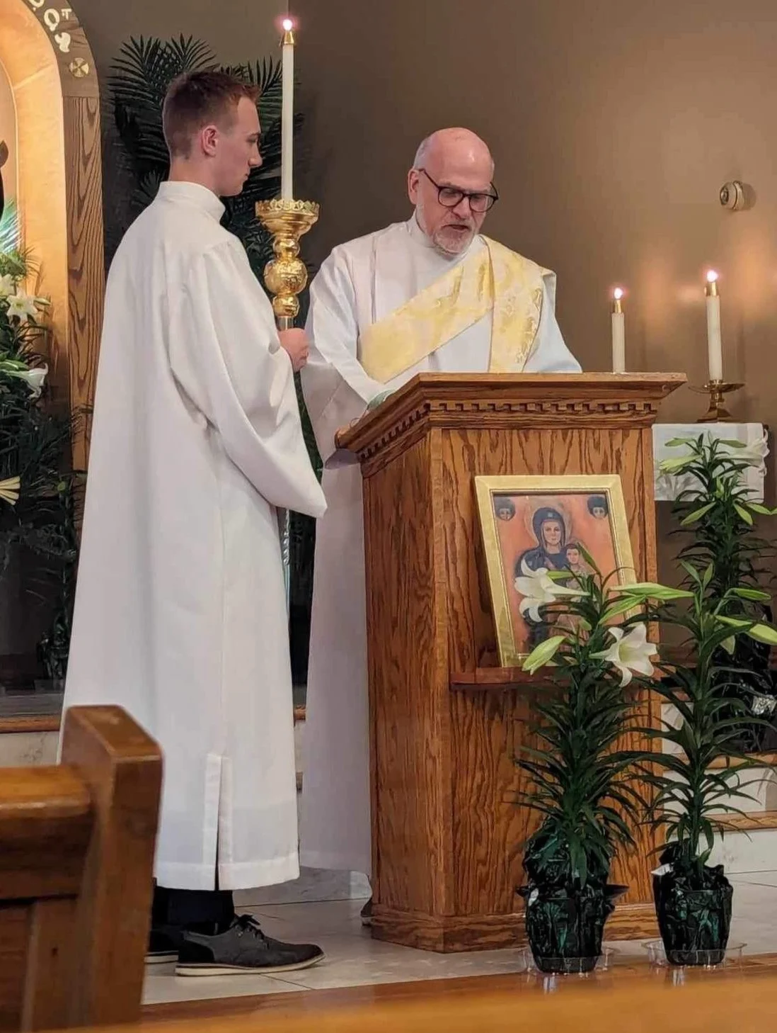 A priest and an altar server stand at a wooden lectern during a church service, with lit candles and religious icons nearby.