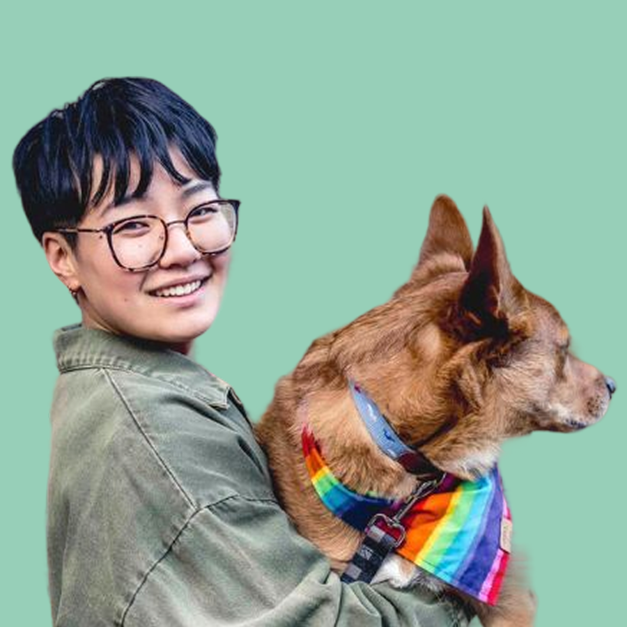 A person with short dark hair, glasses, and a khaki jacket smiling while holding a dog wearing a rainbow pride bandana against a mint green background.