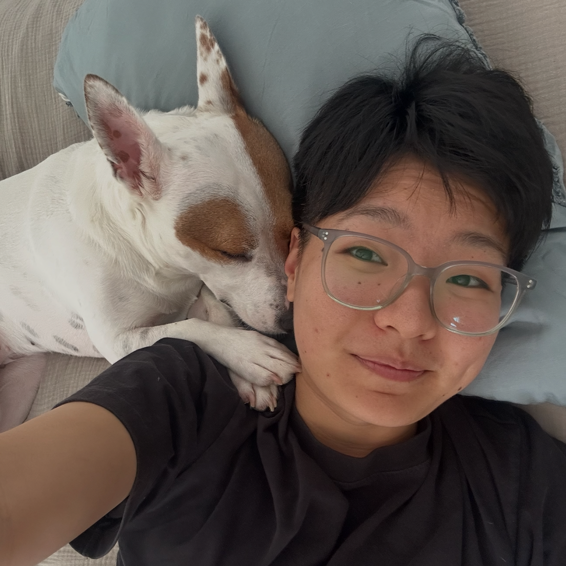 A person with short dark hair and glasses lying on a bed, smiling, with a white and brown dog sleeping next to their face, resting on a pillow.