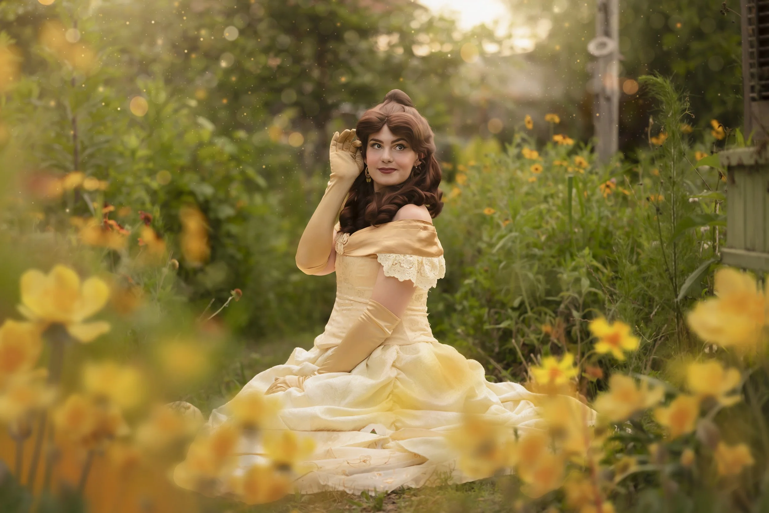 A woman in a vintage, off-shoulder, cream-colored gown with lace details, sitting on the ground surrounded by yellow flowers in a garden during sunset.