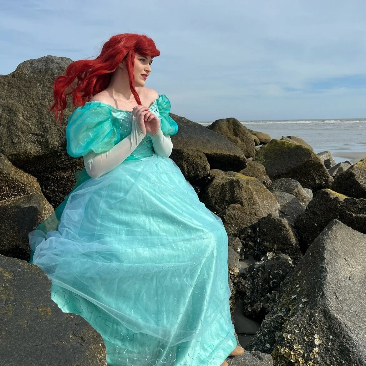 A woman with long red hair in a blue princess dress sitting on rocks by the ocean.