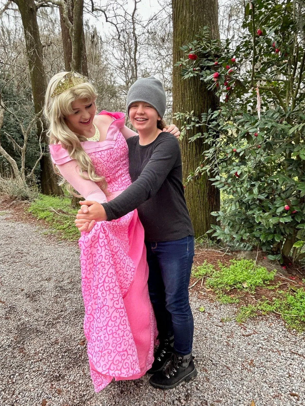 A young girl wearing a grey beanie and a black long-sleeve shirt is hugging a woman dressed as a pink princess with a tiara, outdoors in a wooded area with trees and bushes.