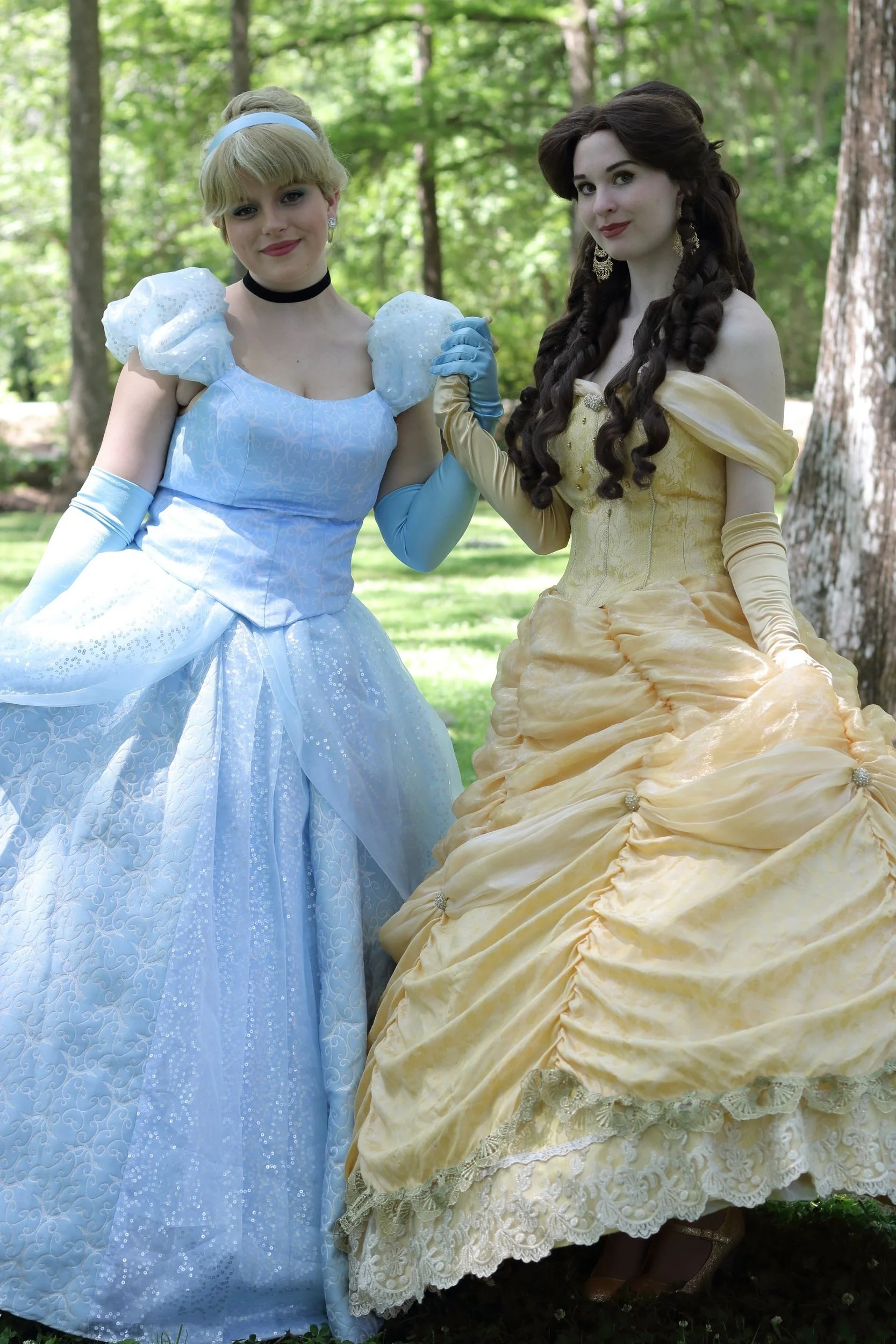 Two women dressed as Disney princesses standing outdoors in a wooded area, holding hands.