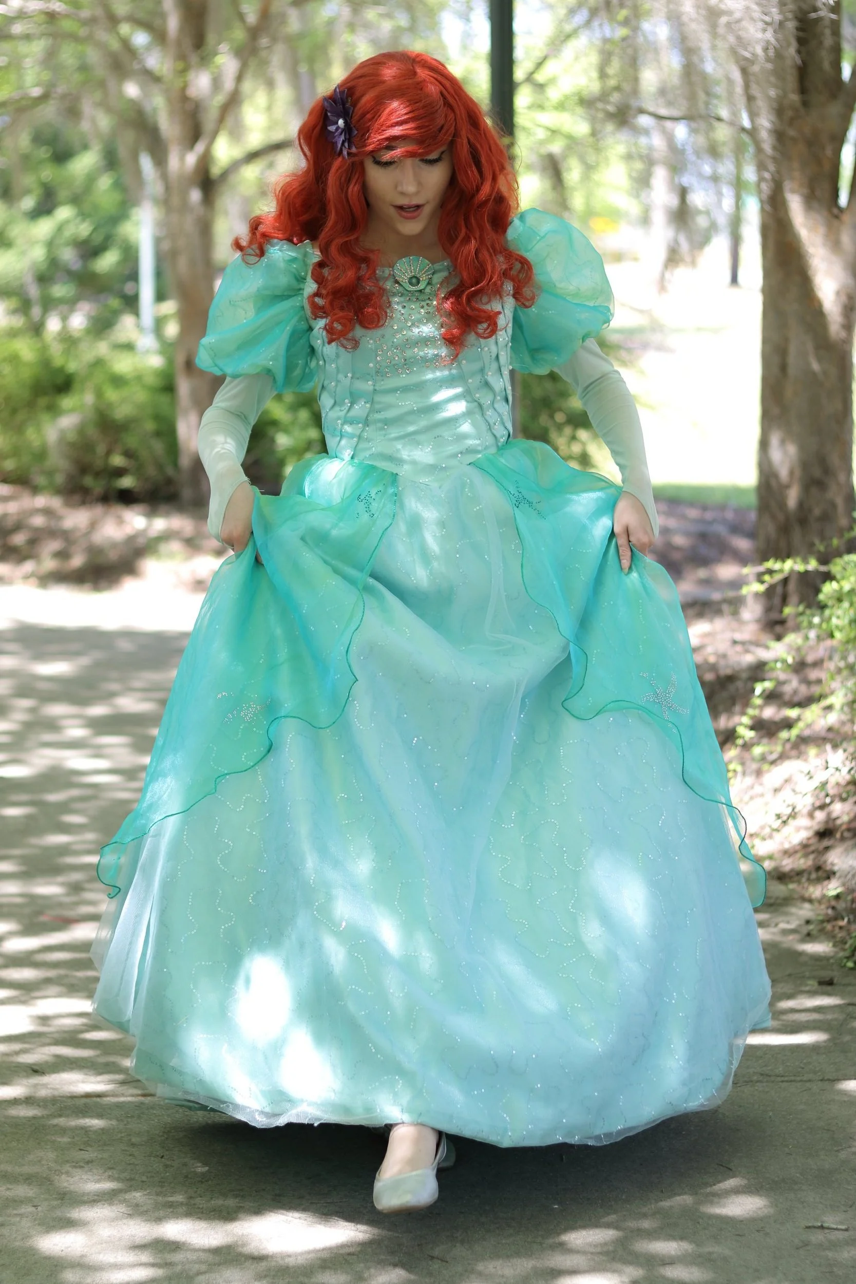 Person dressed as Disney Princess Ariel walking outdoors on a shaded pathway, holding the sides of her turquoise dress.