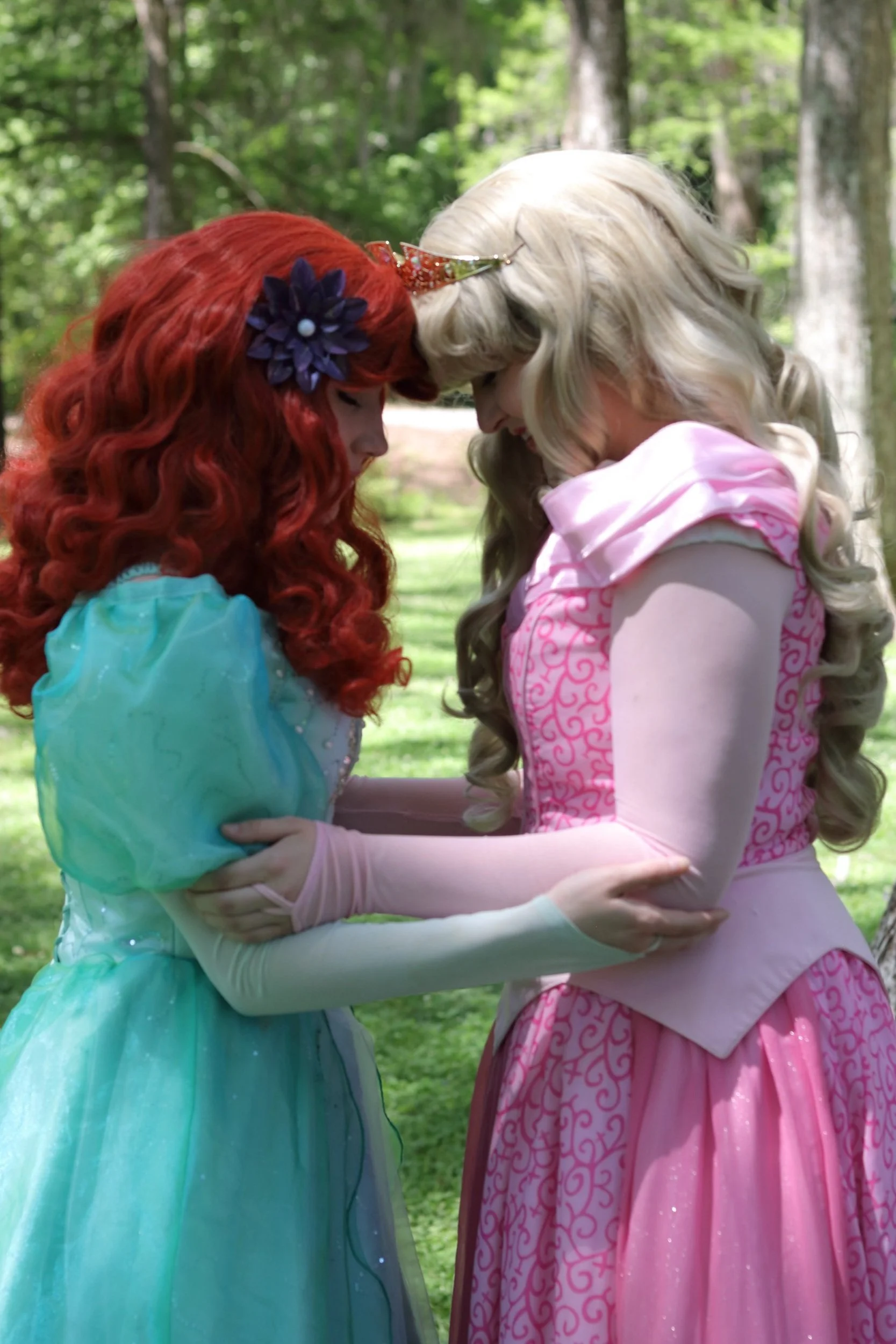 Two women dressed as fairy tale princesses, one with red hair and a blue dress, and the other with blonde hair and a pink dress, are touching foreheads and holding each other's arms in a wooded outdoor setting.
