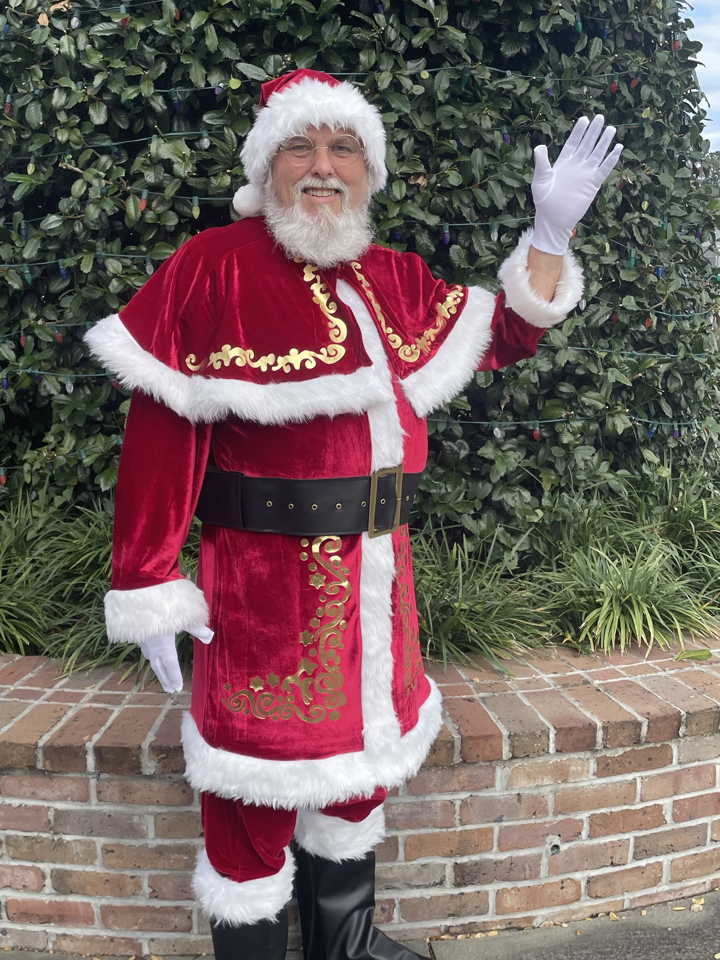 A man dressed as Santa Claus waving and smiling, standing outdoors in front of green foliage.