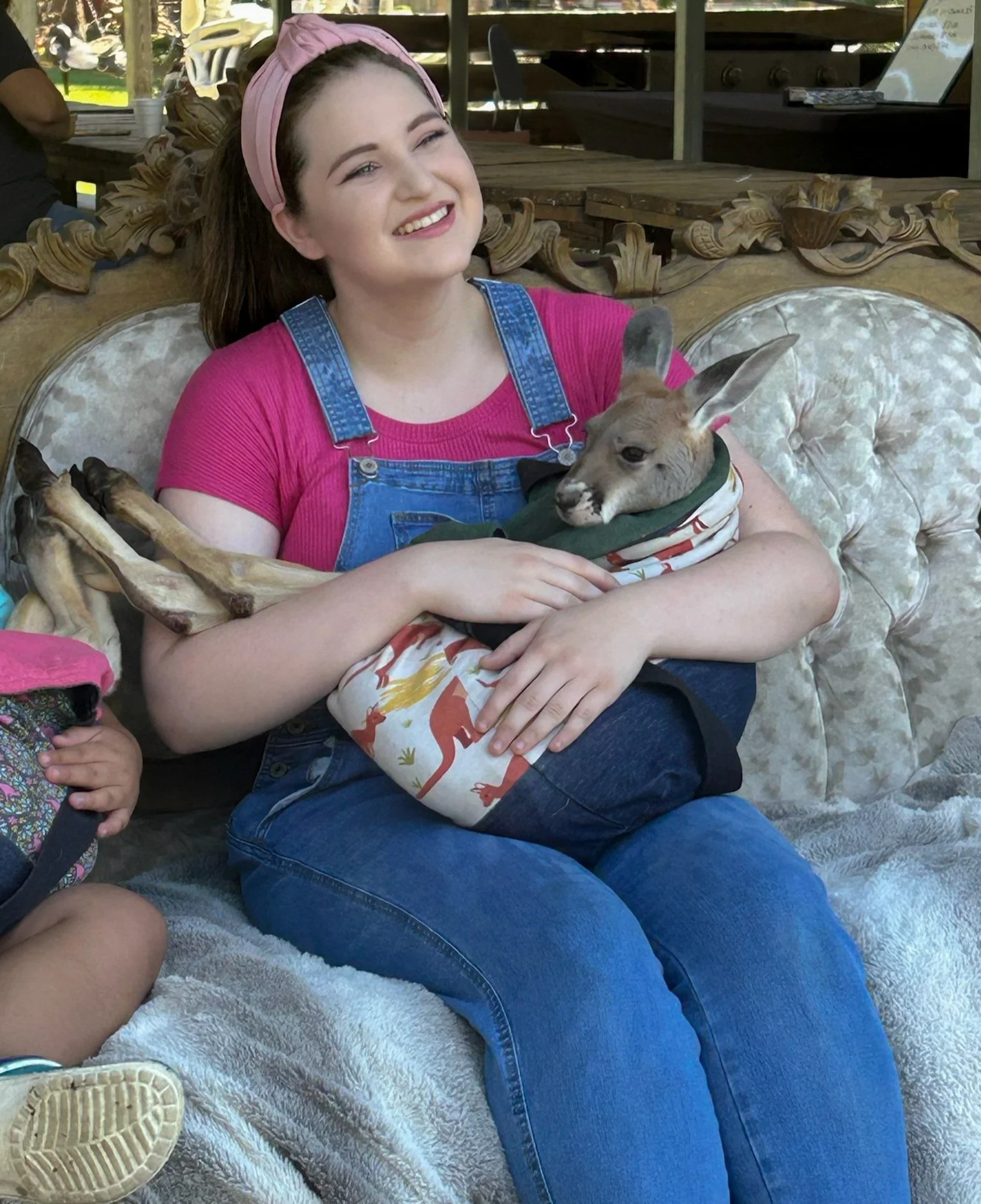 A young woman with brown hair, wearing a pink headband, a pink shirt, and denim overalls, sitting on a beige tufted sofa. She is smiling and holding a kangaroo in her arms.