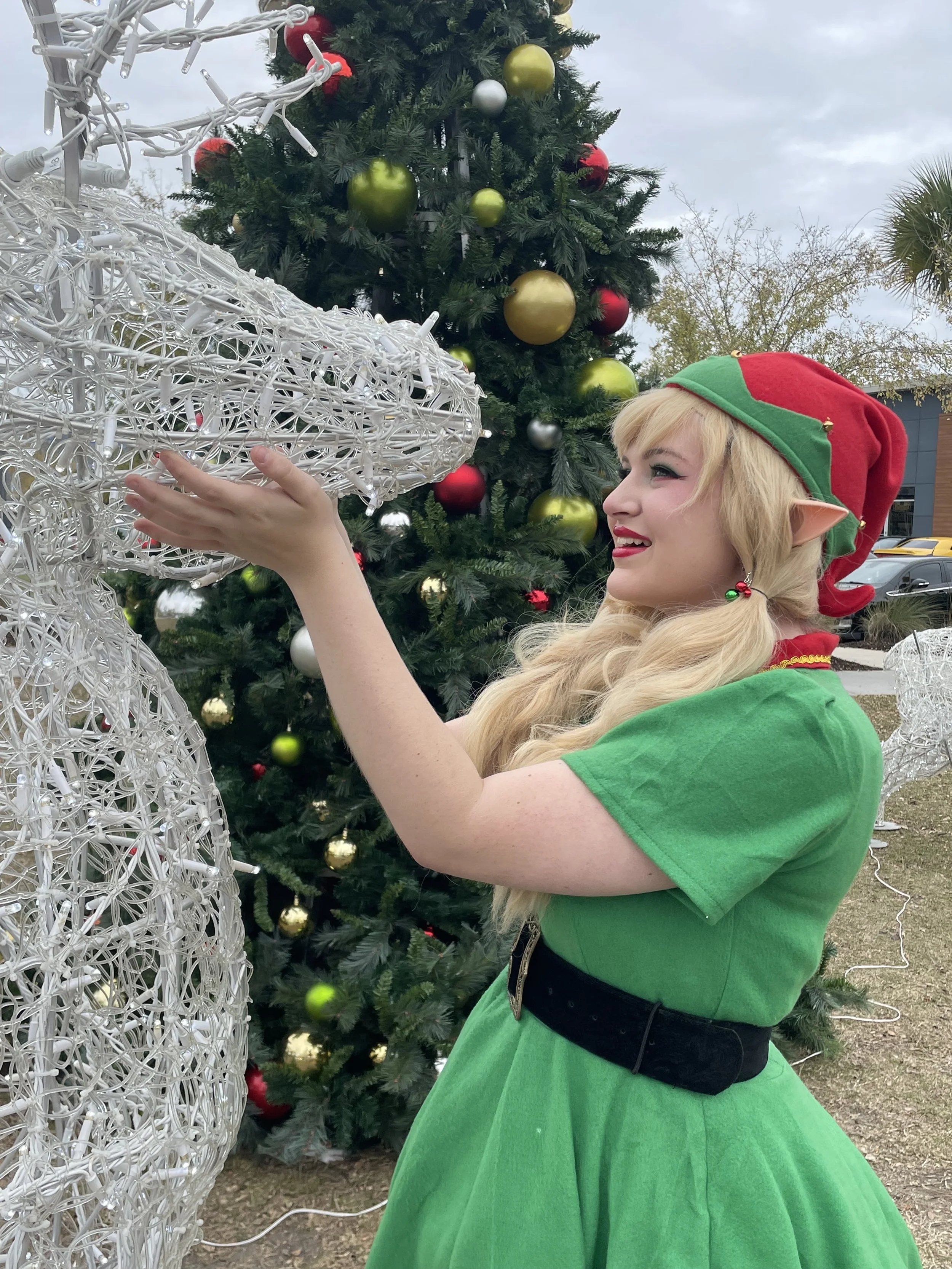 A woman dressed as an elf with long blonde hair, green dress, red and green elf hat, and elf ears, decorating a white light reindeer figure in front of a decorated Christmas tree outdoors.