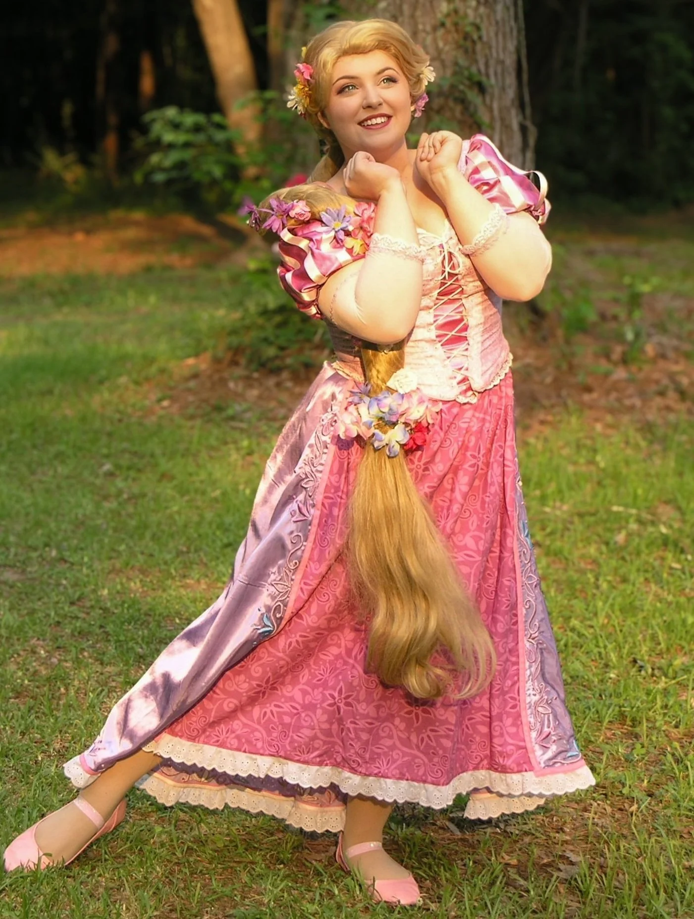 A woman dressed as Rapunzel with long, blonde hair and a pink dress standing outdoors in a wooded area, holding her long hair with flowers woven into it.