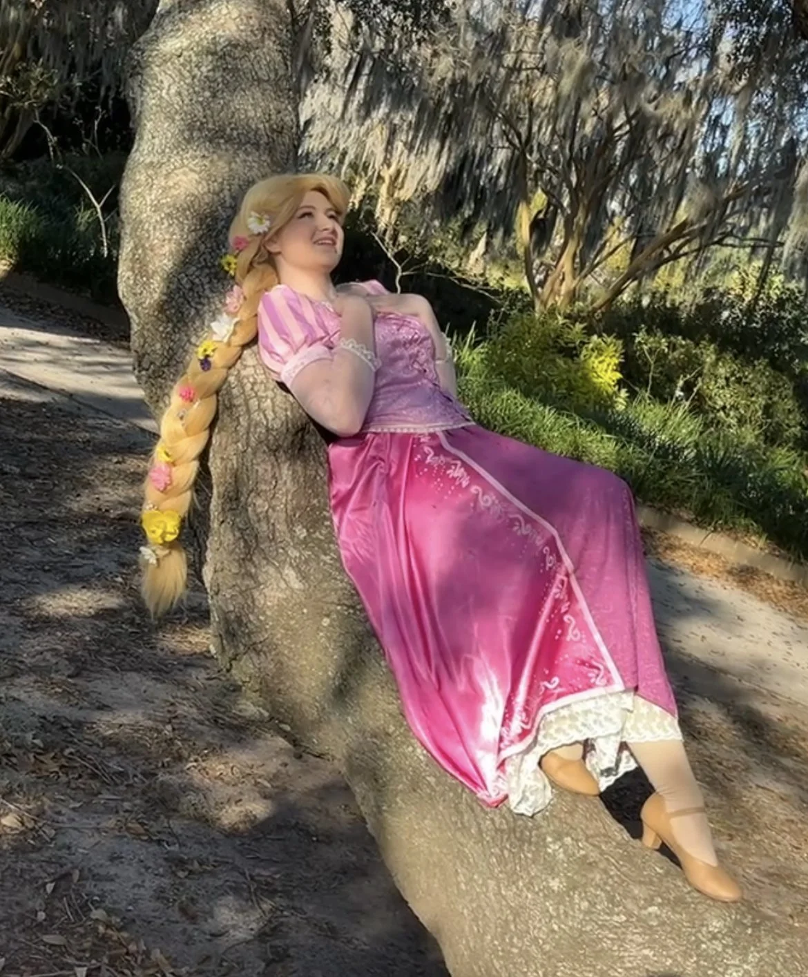 A woman dressed as Rapunzel from fairy tales, wearing a pink and purple dress, lying on a tree branch outdoors with greenery in the background.