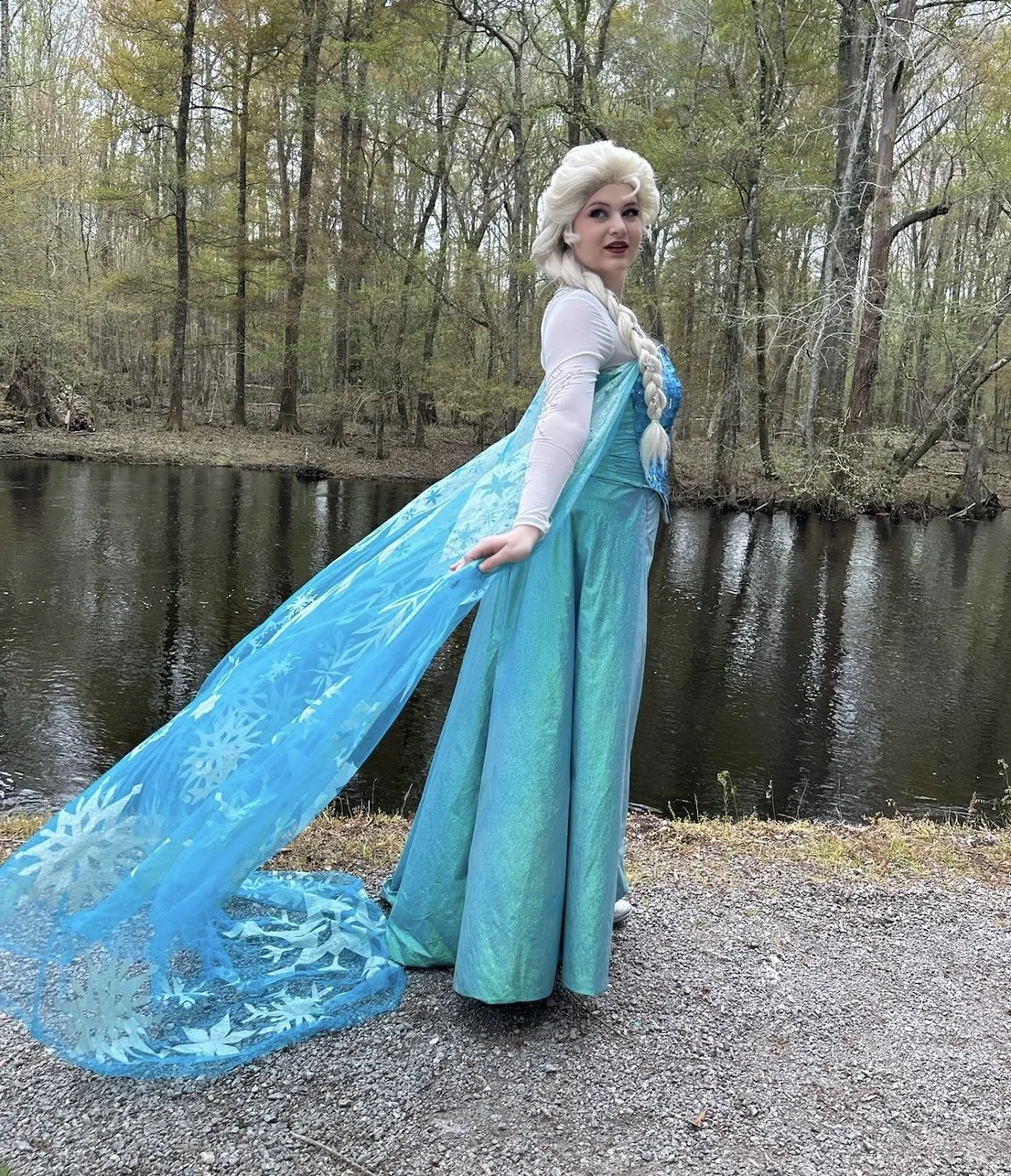A woman dressed as Elsa from Frozen standing outdoors near a river in a wooded area. She is wearing a blue gown and a flowing blue cape with snowflake patterns, and has platinum blonde hair styled in a braid.