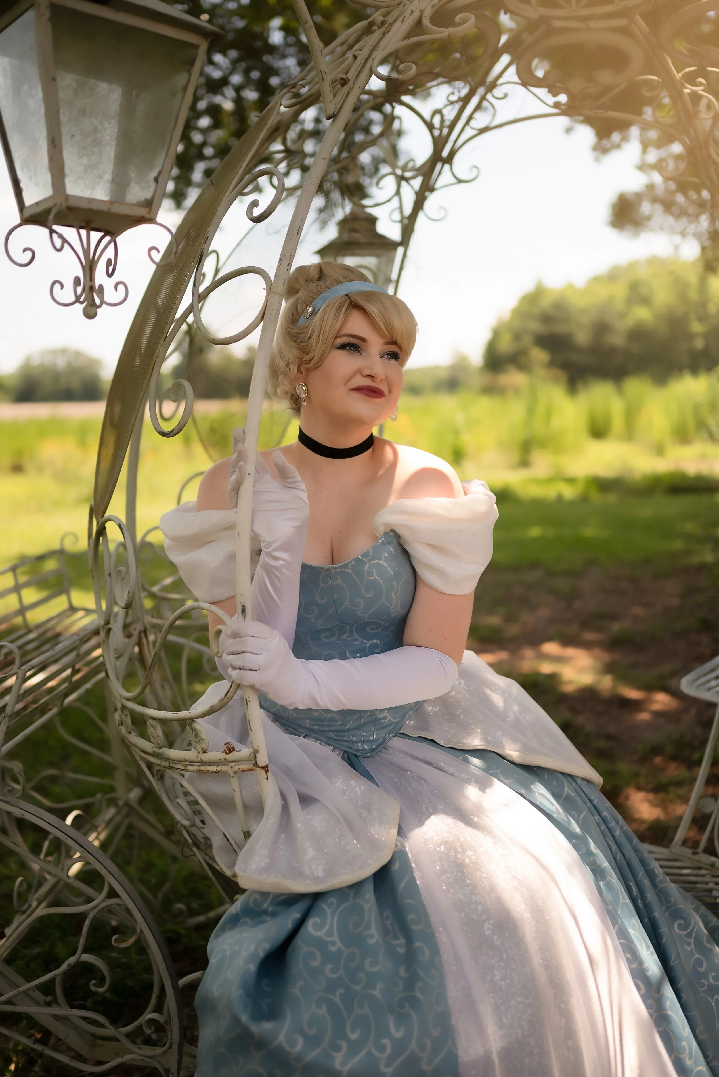 A woman dressed as Cinderella sitting on a decorative white metal carriage outdoors under a structure with lanterns, with greenery and trees in the background.