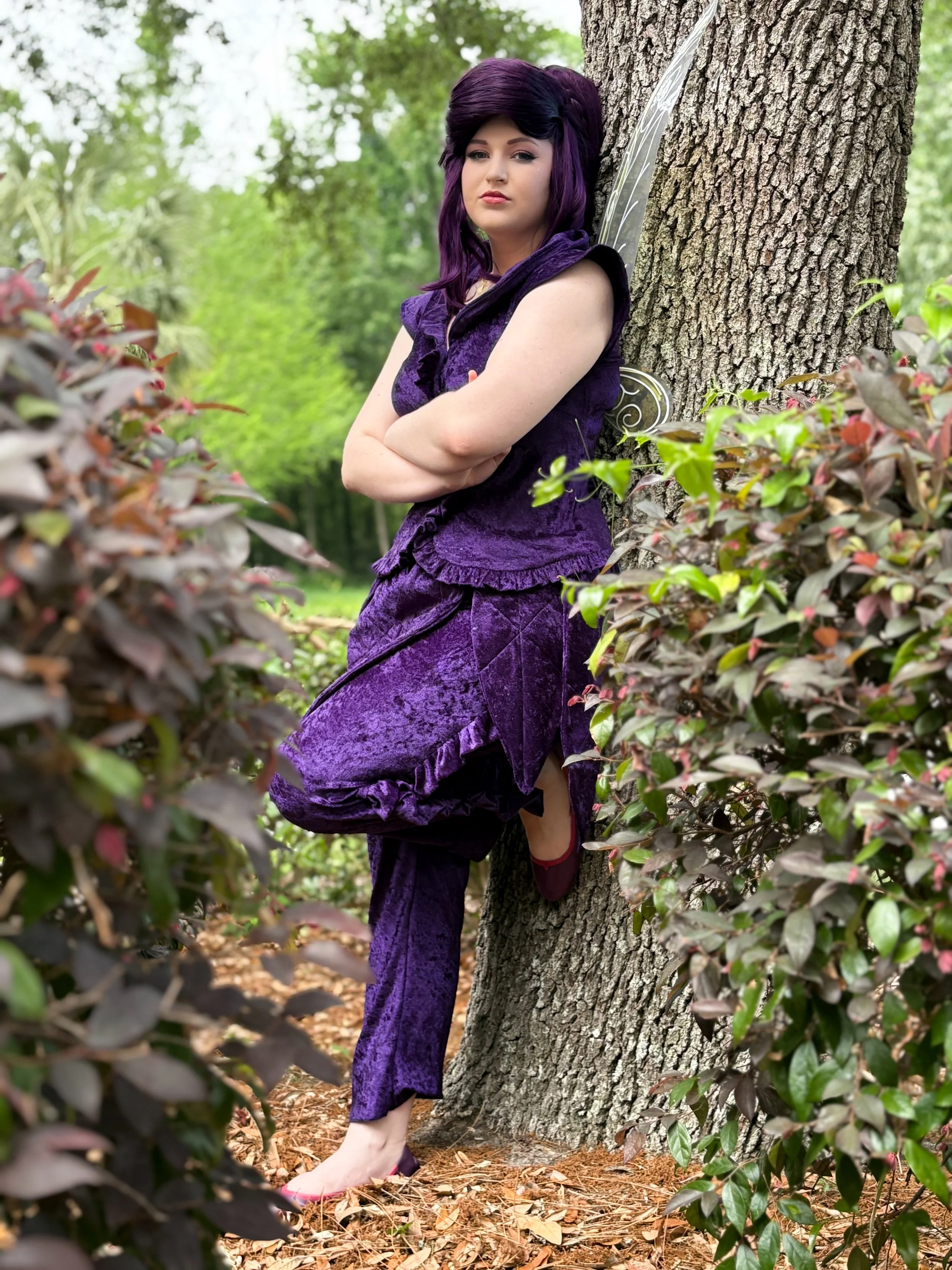 A woman with purple hair and fairy wings leaning against a tree in a purple velvet outfit in a wooded area.